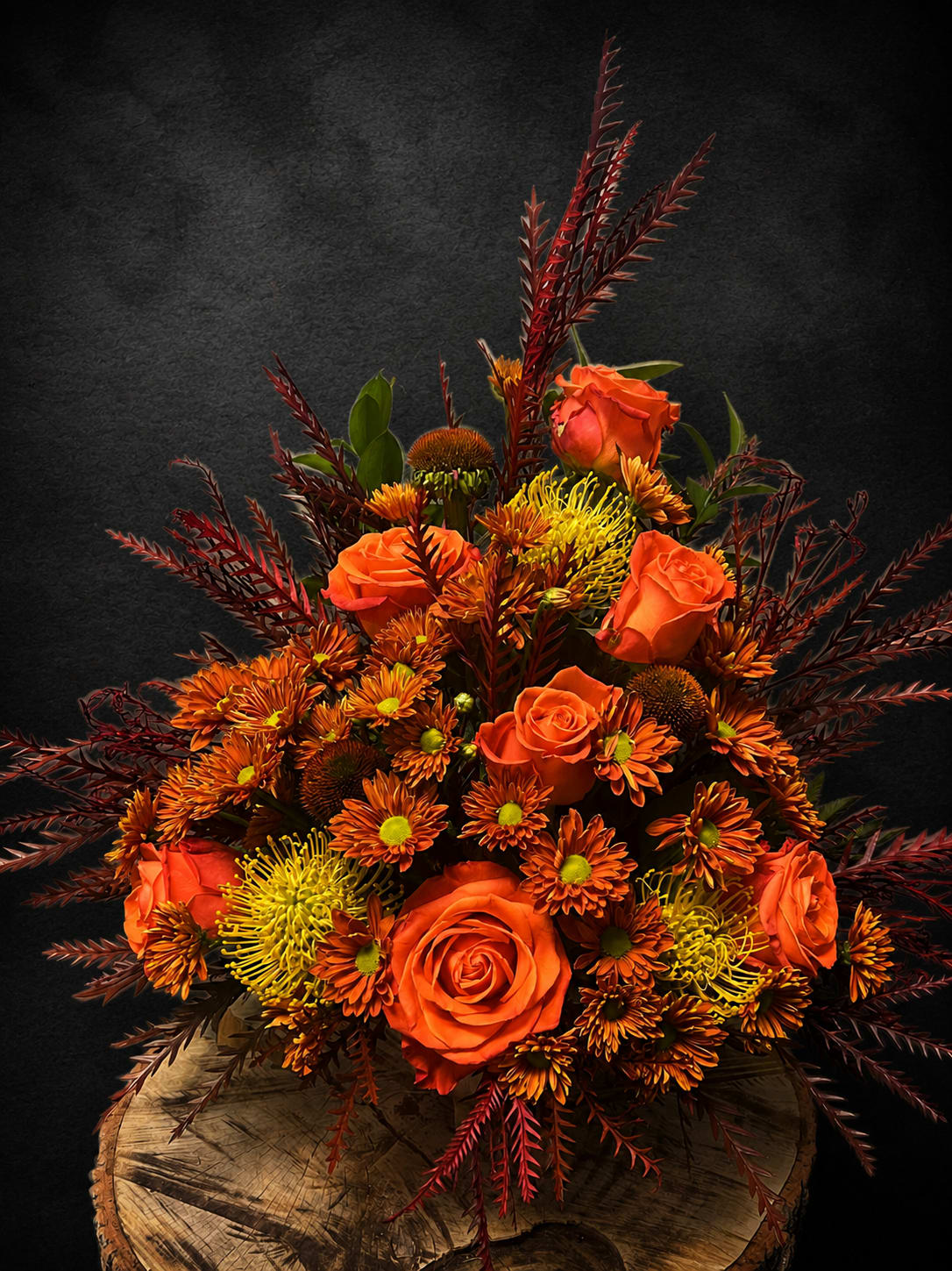 Fall Reflection - Bright, warm and full of autumnal texture, this fall flower arrangement is made to make someone feel truly seen. The flower arrangement features vivid orange roses and clusters of yellow spider mums and chrysanthemums arranged on a rustic wooden base in a low basket - perfect for an anniversary centerpiece, a birthday surprise, or a thoughtful Thanksgiving or housewarming gift that reads as cozy and celebratory.  Tactile and dynamic, the design pairs dense, rounded masses of carnations, daisies, roses and chrysanthemums in yellow, orange, red and mixed tones with tall burgundy, feathery foliage that radiates upward for dramatic height and movement. The low, grounded profile of the basket keeps the composition intimate while the spiky accents give it seasonal energy; the wooden base adds a rustic, homey finish that complements autumn tables and cozy living spaces.  Whether you're sending a warm "thinking of you," a cheerful thank-you, an "I'm sorry," or a loving anniversary or retirement message, this arrangement feels like a gift that comforts and delights. The compact, full design is easy to display on a table, mantel or entryway - order now for same-season delivery during fall holidays or anytime you want to brighten someone's day.
