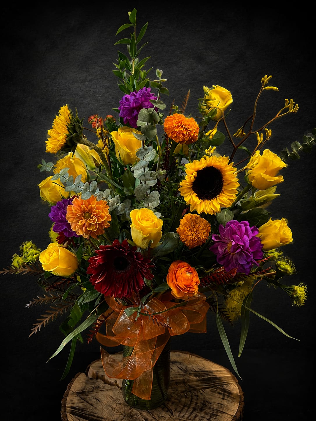 Happy Day Bouquet  - Sunflower-bright and full of cheer, this tall flower arrangement puts yellow roses and sunflowers center stage for an immediate lift-perfect as an anniversary bouquet, a get-well pick-me-up, or a bold housewarming surprise. The Happy Day Bouquet also reads beautifully for autumn and Thanksgiving displays while working just as well year-round for birthdays, thank-you gestures, thinking-of-you moments, retirement celebrations, or a sincere "I'm sorry."  Densely layered for a dramatic upright silhouette, the mix includes yellow roses and sunflowers paired with purple and orange dahlias and gerbera daisies, plus deep red accents and seasonal filler greenery for texture. Arranged in a clear glass vase and finished with a sheer orange ribbon, the composition is tall, abundant, and colorful-designed to feel festive and celebratory from every angle.  This bouquet makes the recipient feel genuinely seen-bright, warm, and uplifted-whether you're saying congrats, honoring Mother's Day or Grandparent's Day, or sending sympathy with heartfelt color. Available in a full, tall size as shown; vase included. For best enjoyment, trim stems, refresh water every two days, and keep out of direct heat or drafts.