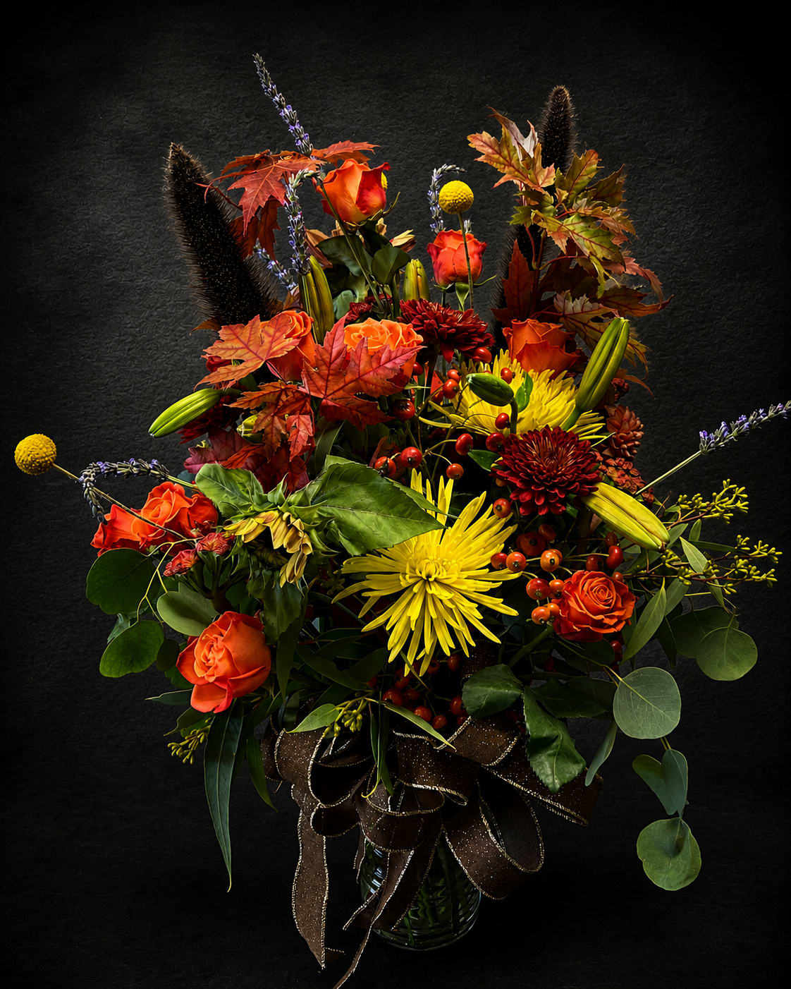 Happy Fall Y’all - A bold harvest bouquet that reads like autumn in full color-orange roses, bright yellow chrysanthemums and marigolds, and deep red accent blooms are layered with seasonal foliage, berry sprays and tall textured stems to create a dramatic birthday bouquet or Thanksgiving centerpiece. This flower arrangement is ideal for anniversary, retirement, or holiday gifting when you want the person you're thinking of to feel embraced by warmth and abundance; it also works beautifully as a get well, thank you, thinking of you, or sympathy gesture that says you care.  Carefully arranged in a clear glass vase and finished with a wide decorative ribbon, the display combines lilies, stock, and other seasonal blooms with dense greenery and maple‑like leaves for a full, vertical silhouette. Tall seed heads and arching lavender‑like stems add height and contrast, while the mix of orange, peach, gold, burgundy and hints of purple gives depth and a rich harvest mood. This large arrangement makes a striking centerpiece for Thanksgiving or any autumn table and arrives ready to display-refresh water daily and recut stems if you need extra longevity for a special occasion.