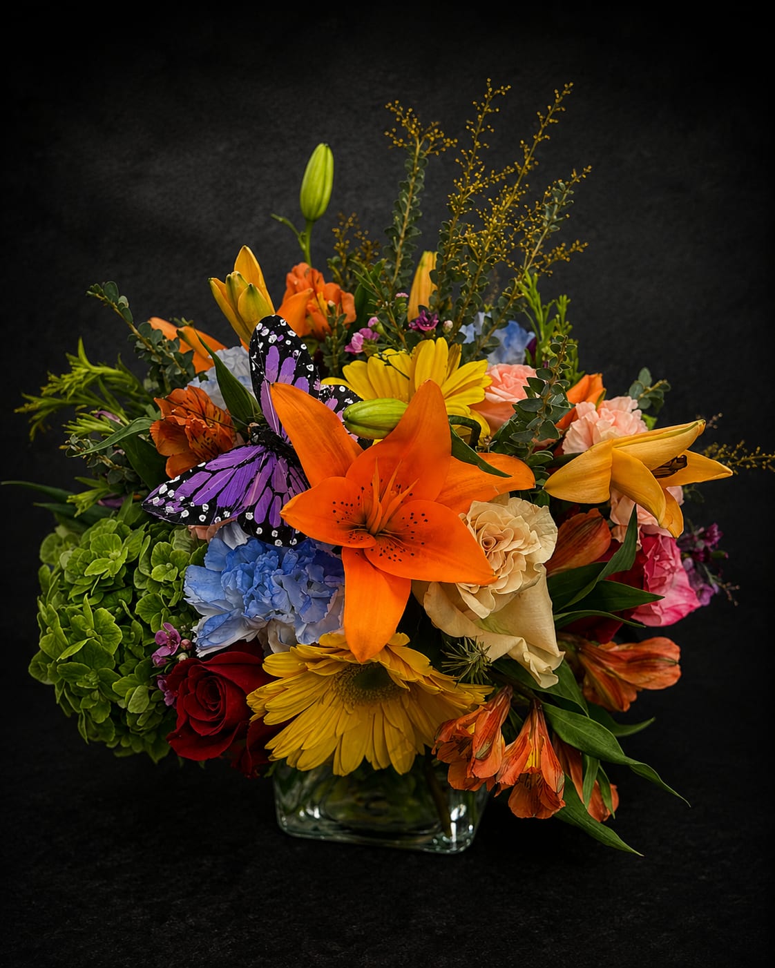 Designer’s Choice  - A vibrant, mixed-color Designer's Choice flower arrangement that makes a joyful anniversary gift or birthday surprise-built to delight the person you're thinking of. Whether you need a cheerful get-well bouquet, a romantic anniversary display, or a bright just-because pick-me-up, this rounded arrangement reads as celebratory and thoughtful. Warm orange lilies and sunny yellow daisy-like blooms catch the eye, while roses in hot pink, blush, cream and mauve add romantic depth; the overall palette spans pink, purple, lavender, orange, yellow, blue, green and white for wide seasonal appeal. 