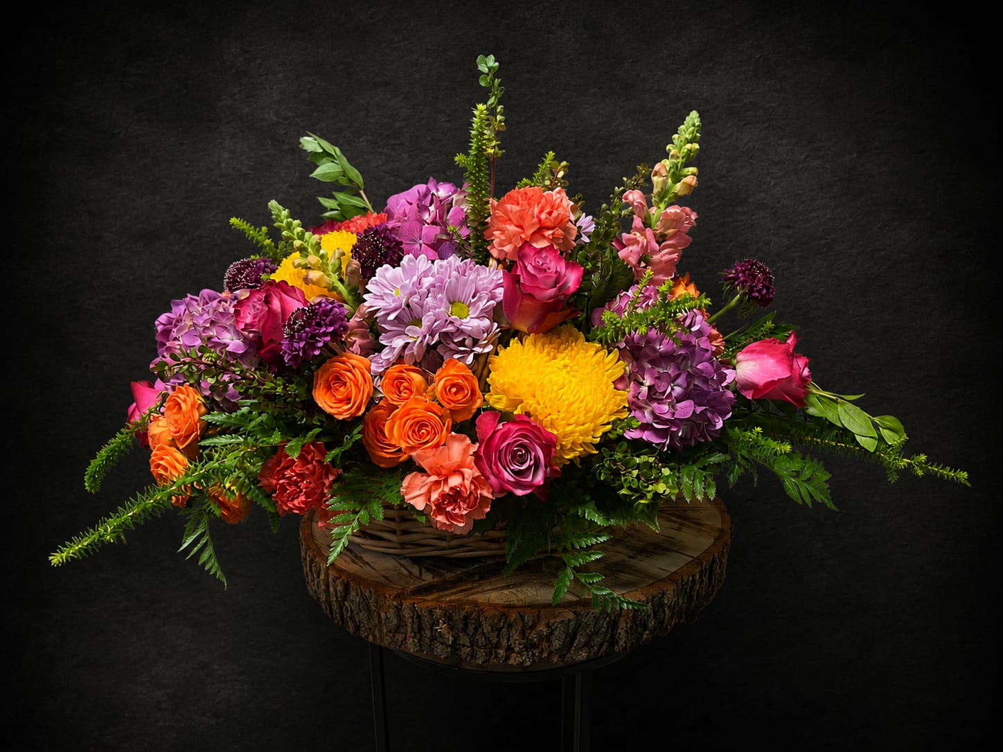 A Special Thank You - Lively pinks, purples, oranges and sunny yellows burst from a rustic woven basket in this garden-style flower arrangement-perfect as a thank you bouquet or for birthdays, anniversaries, housewarmings, and just-because surprises. The rounded, abundant shape reads as joyful and celebratory, the kind of gift that makes the person you're thinking of feel truly seen and appreciated.  A dense mix of garden roses, classic roses, spray roses, carnations, chrysanthemums, hydrangea-like clusters, stock, snapdragons and cheerful daisies creates rich, layered texture from low, full blooms to taller snapdragon spikes that add vertical interest. The arrangement sits on a wood slab within a wicker basket for a handcrafted, natural presentation that plays well in casual or formal settings and complements springtime gatherings as easily as everyday décor.  Thoughtfully designed to convey gratitude, sympathy, congratulations or get-well wishes, this piece suits a wide range of occasions-Thank You, Thinking of You, I'm Sorry, Sympathy and Funeral, or a bright pick-me-up for a new home. Size and fullness make it a statement centerpiece for a table or entryway; simple care is keeping the vase or basket liner topped with fresh water and trimming stems if re-arranged into a vase.