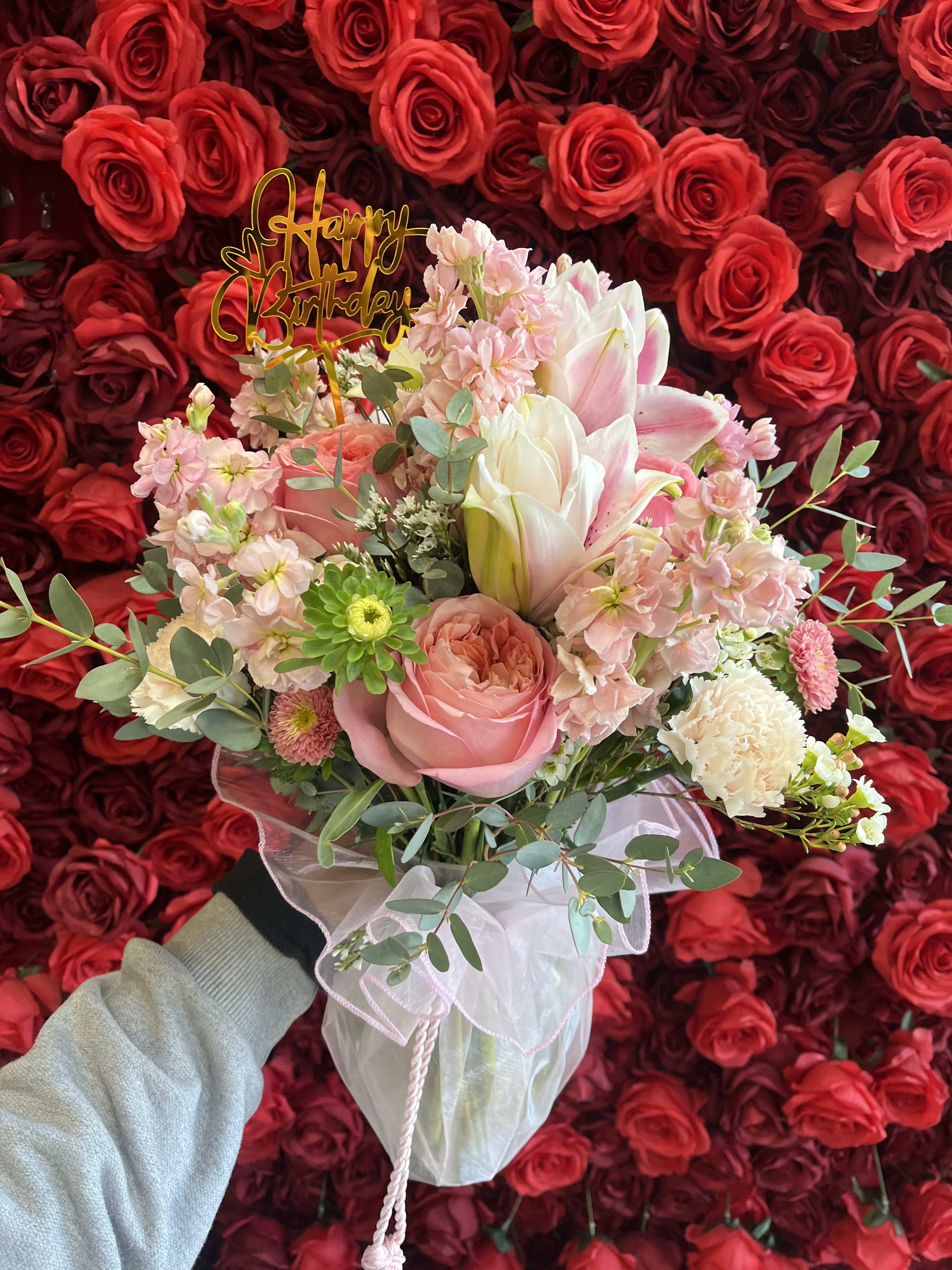 Pink and white bouquet with a birthday topper against a wall of red roses