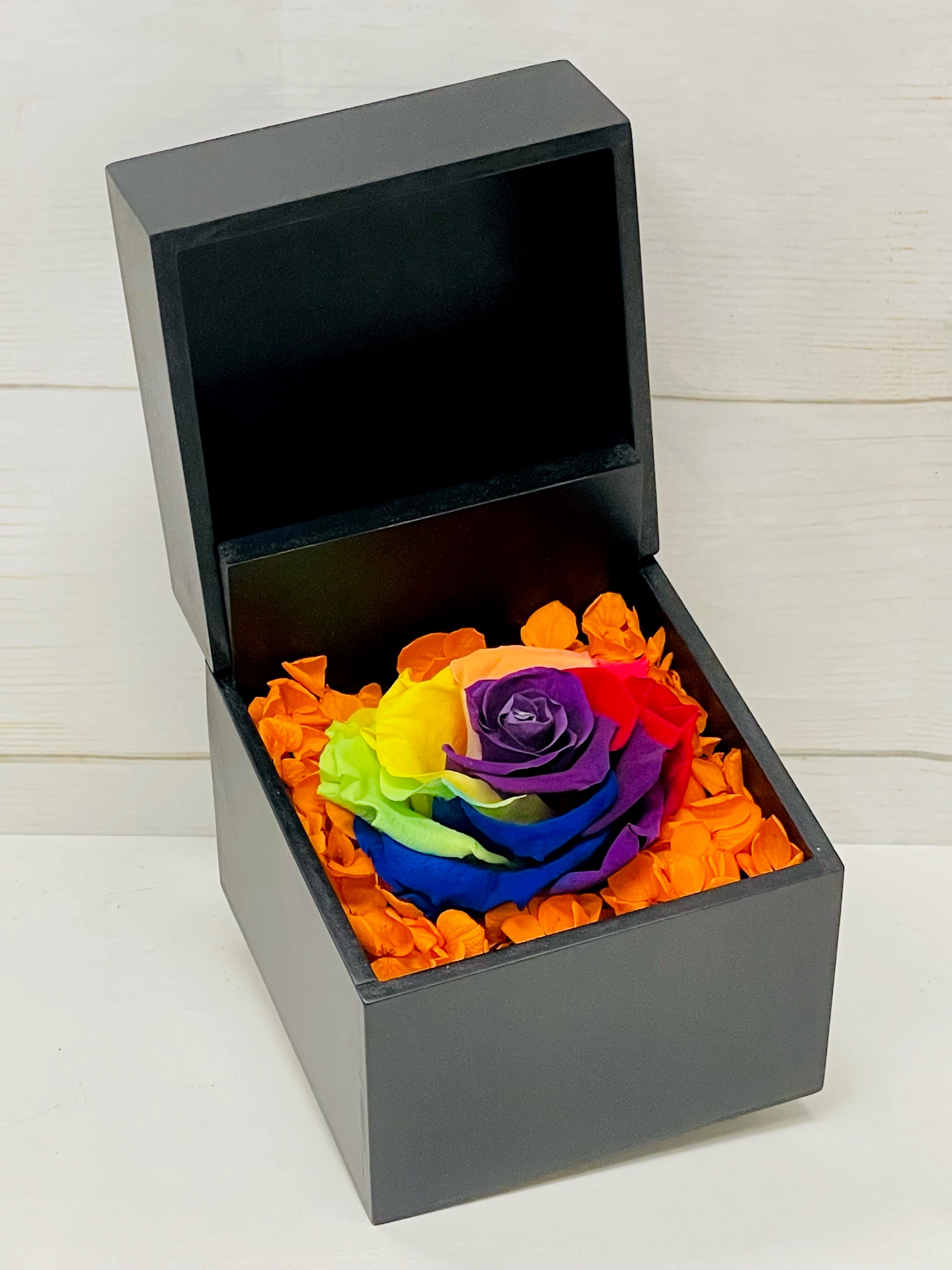 Black gift box holding a rainbow-colored rose surrounded by bright orange petals