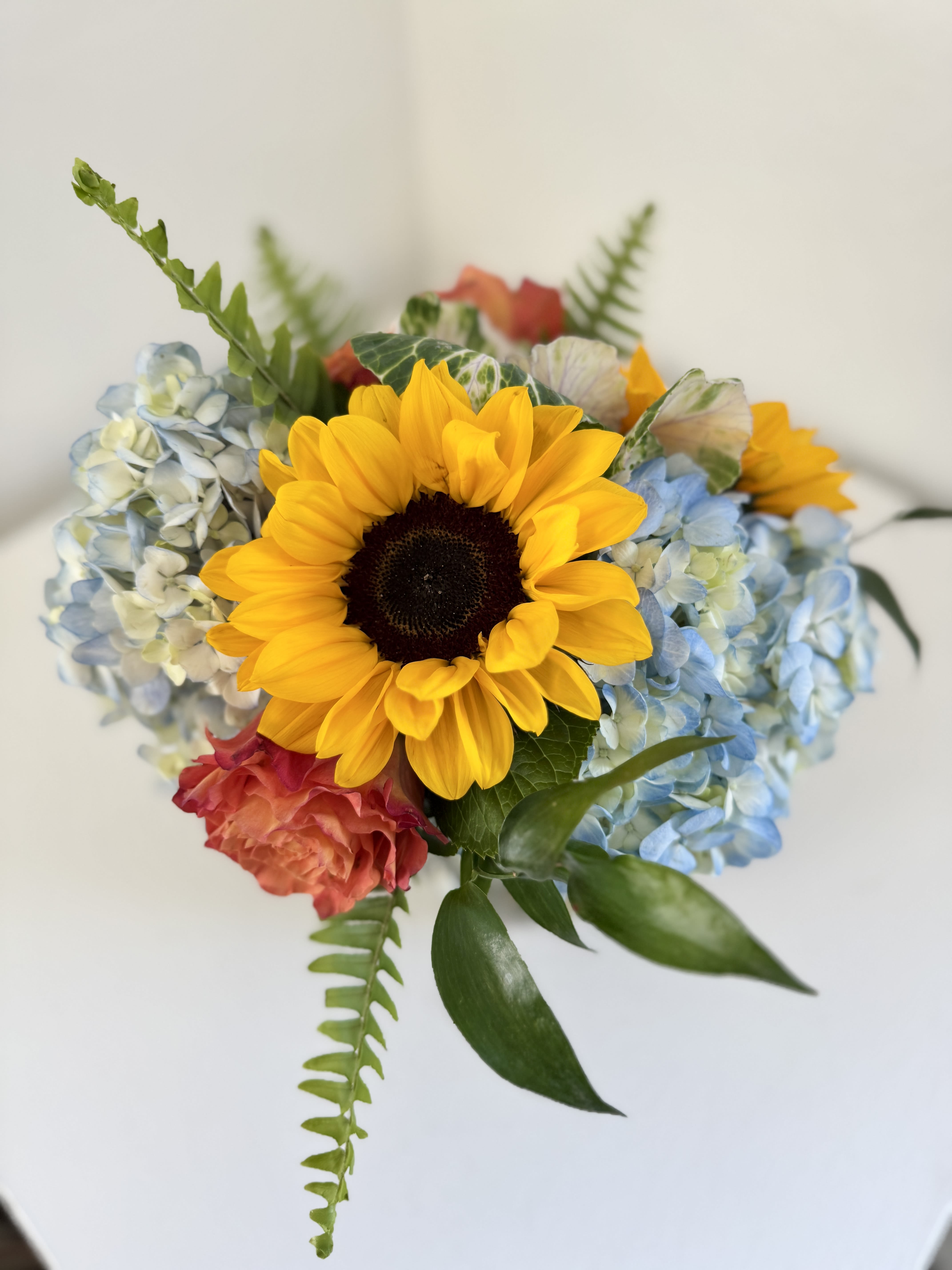 Compact bouquet with a large yellow sunflower, blue hydrangeas, and coral roses.