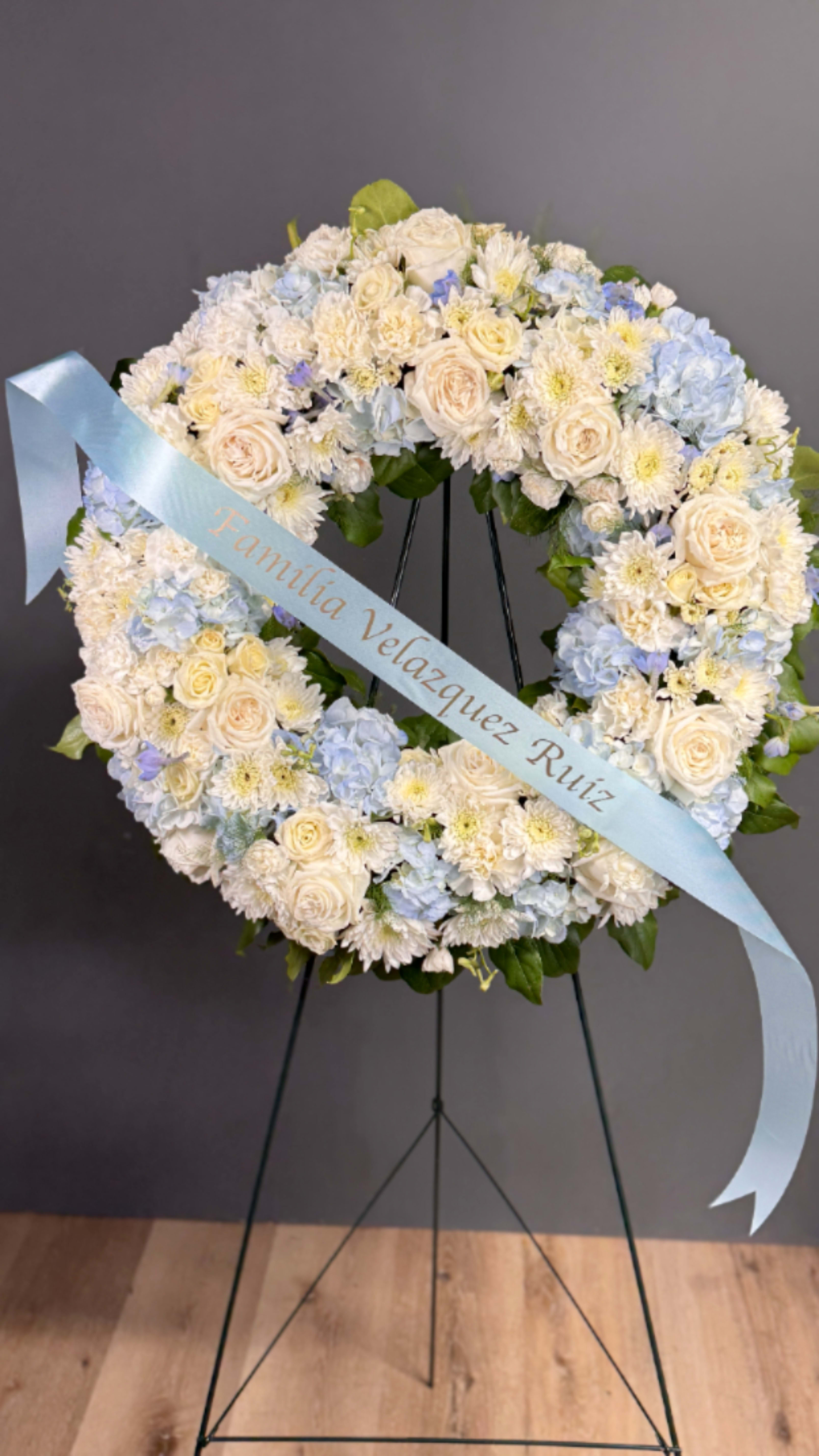 Standing wreath of white and pale blue flowers with a light blue message ribbon on an easel.