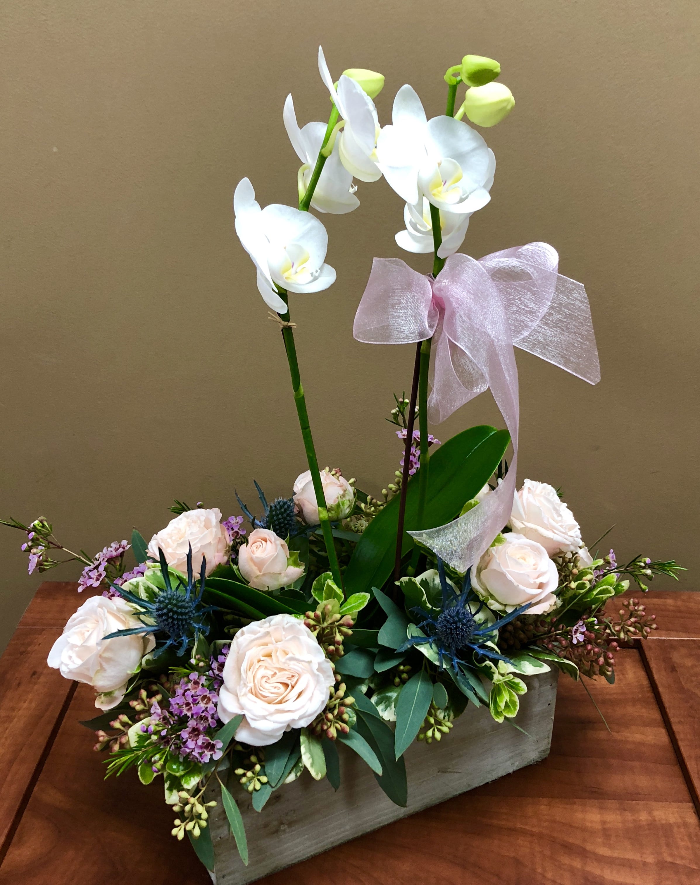 White orchid stems with blush roses and blue accents in a wooden box with a pink bow.