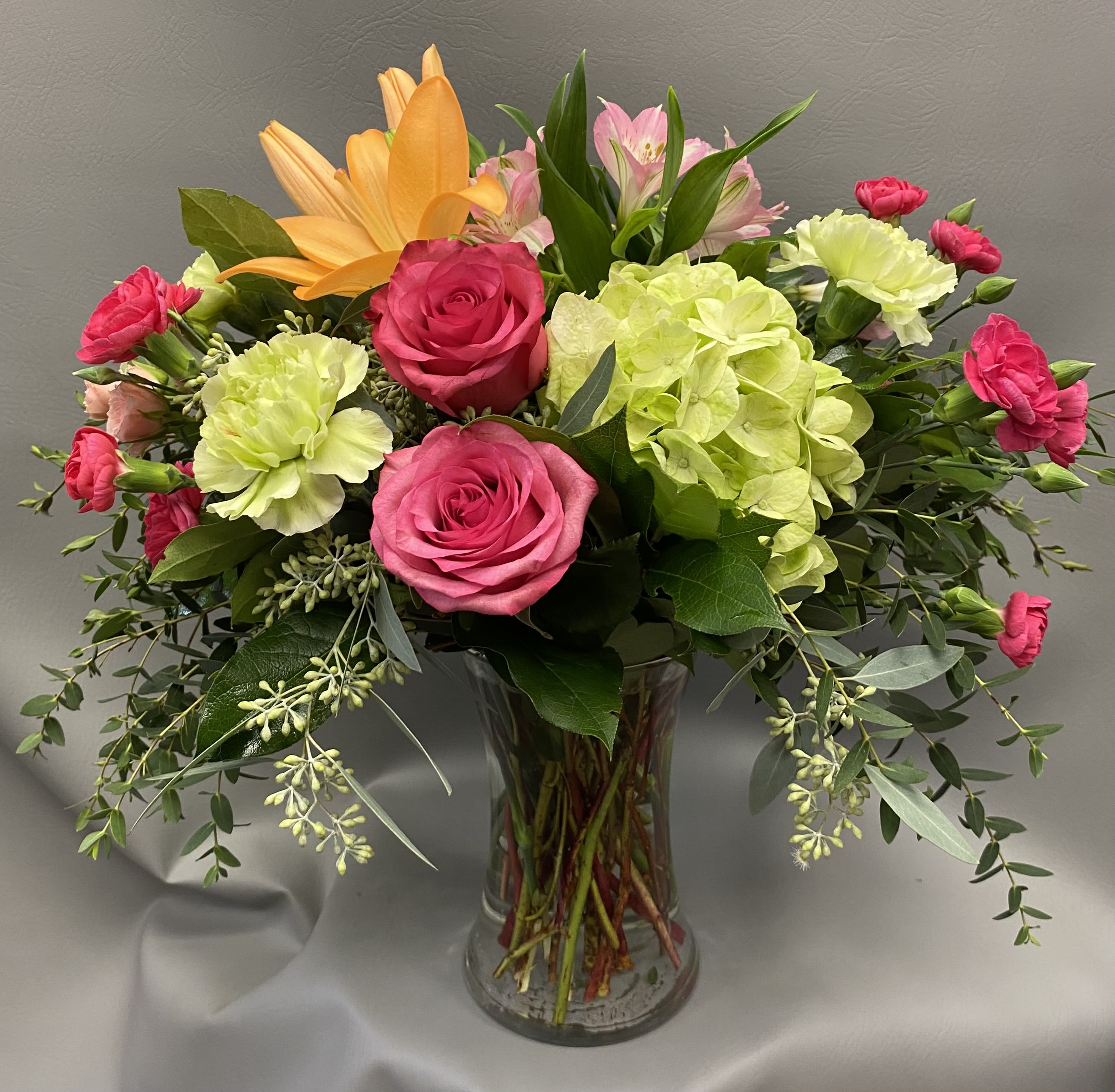 Mixed bouquet of pink roses, green hydrangea, and an orange lily in a clear glass vase