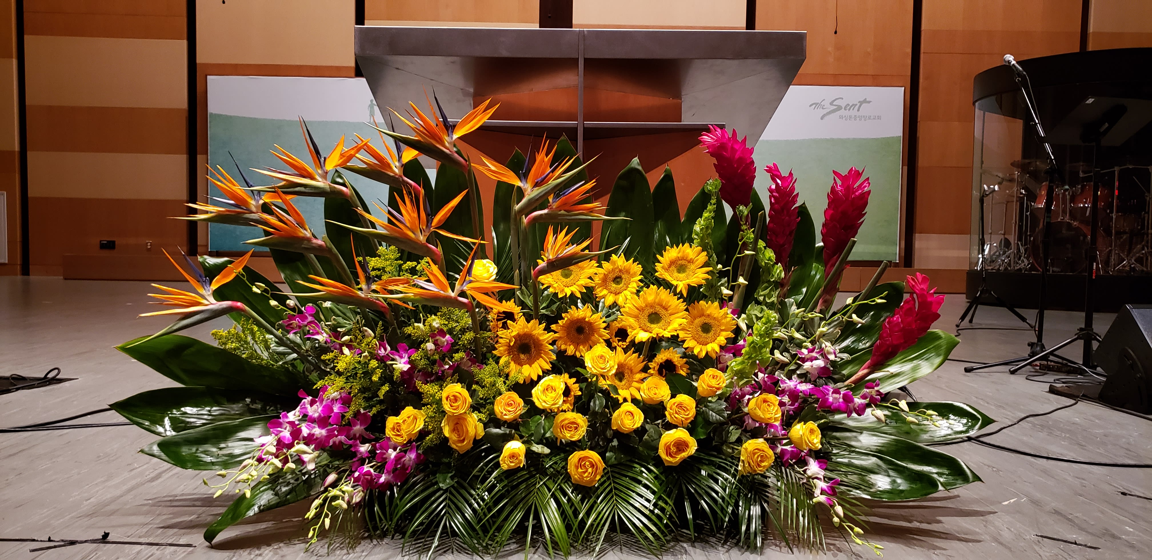 Large tropical stage arrangement with yellow sunflowers, roses, birds of paradise, orchids, and red ginger