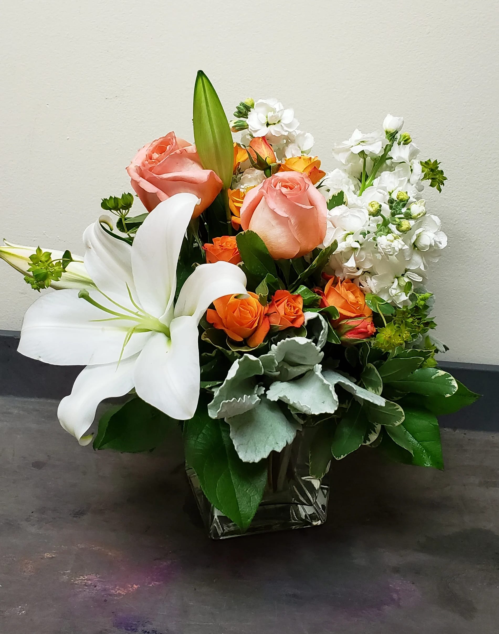 Compact arrangement of white lilies, peach roses, and orange spray roses in a square glass vase