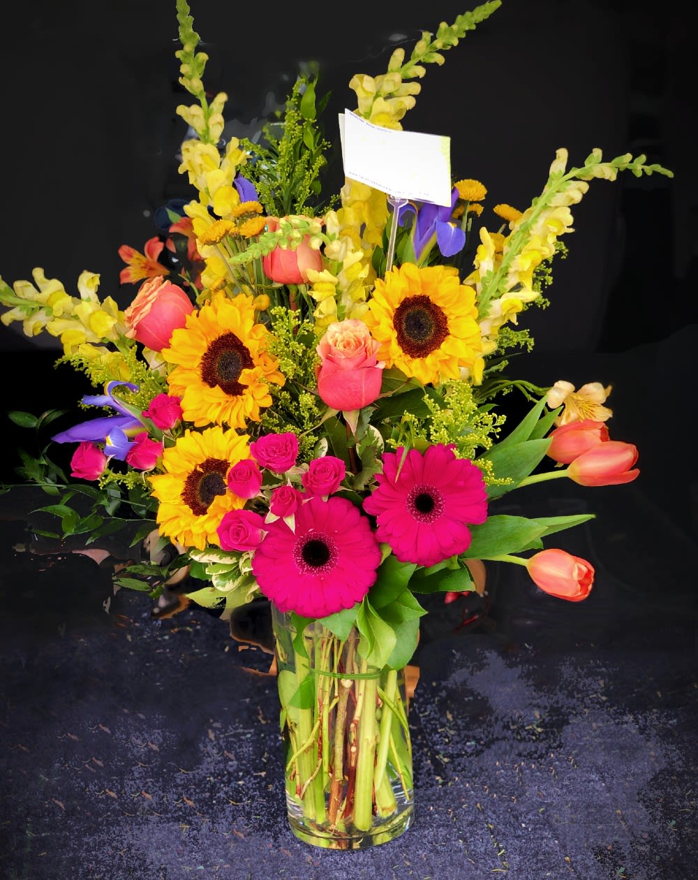 Mixed bouquet with sunflowers, pink gerberas, roses, and tulips in a glass vase