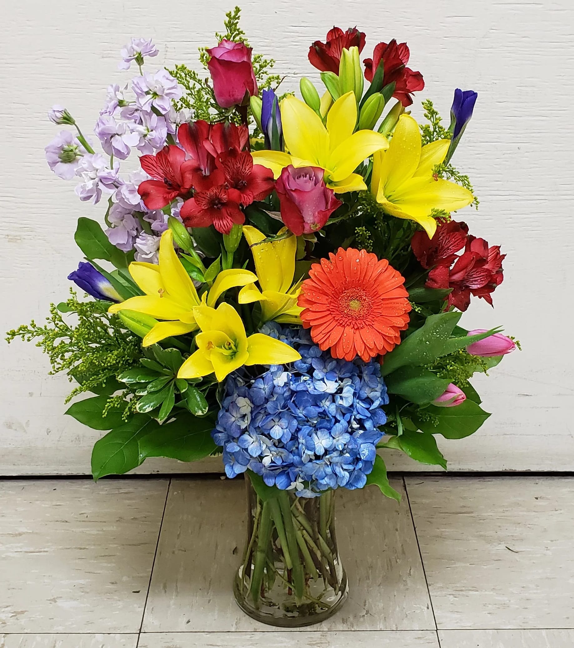 Colorful mixed bouquet in a clear glass vase with lilies, roses, hydrangea, and gerbera daisy