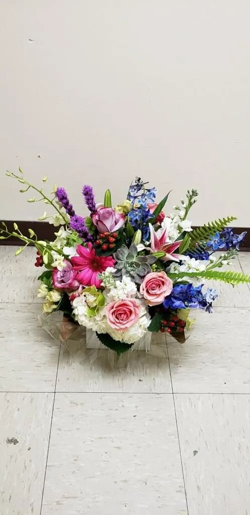 Low mixed arrangement of pink roses, lilies, gerbera daisy, blue delphinium, orchids, and succulents.