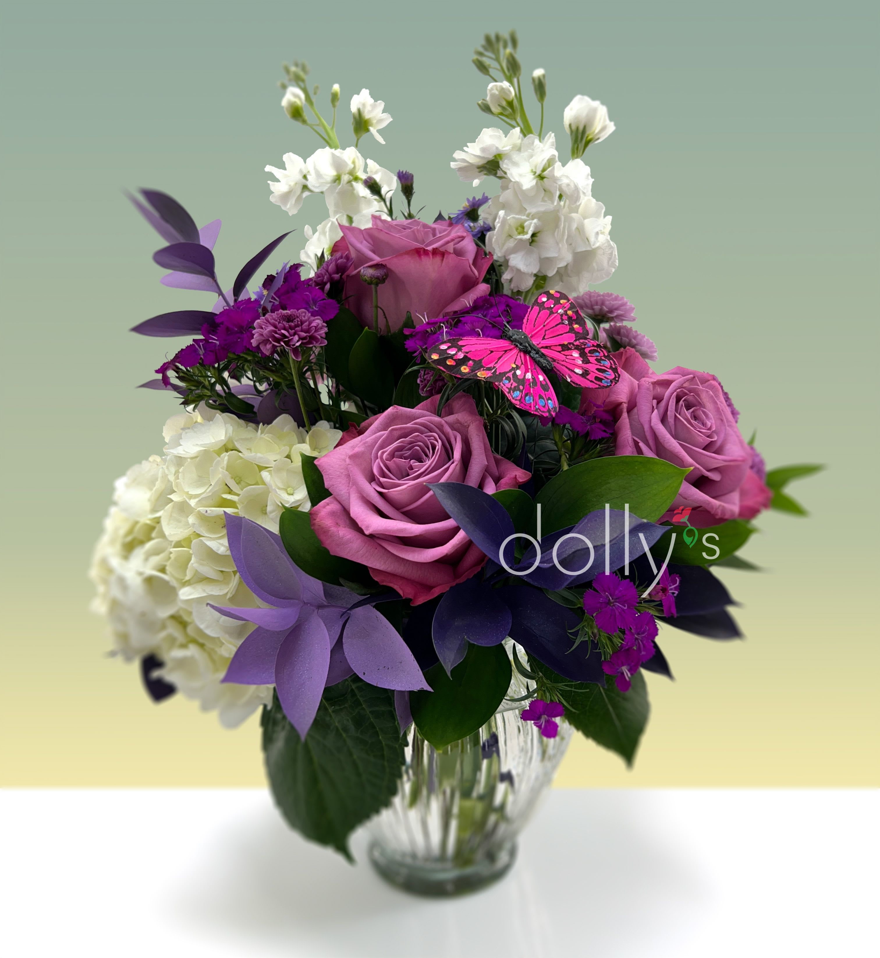 Big Hug - Soft lavender roses and lush white‑green hydrangeas create the first impression of this flower arrangement, a romantic, cheerful choice for a birthday, anniversary, or Mother's Day bouquet. The mix reads as a big hug for the person you're thinking of-gentle pink lilies and magenta dianthus lend warmth while decorative butterfly picks and purple artificial foliage add a whimsical, contemporary touch perfect for Valentine's Day or an everyday pick‑me‑up.  Densely grouped for a full, rounded silhouette, the design layers lavender and plum roses with clusters of deep purple chrysanthemums, violet asters, white stock, and rich purple carnations for texture and contrast. Unopened lily buds and sprays of green tones soften the palette of purple, pink, white, and green. Arranged in a clear ribbed glass vase, the composition measures as a generous centerpiece and arrives ready to display with the decorative butterfly accents in place.