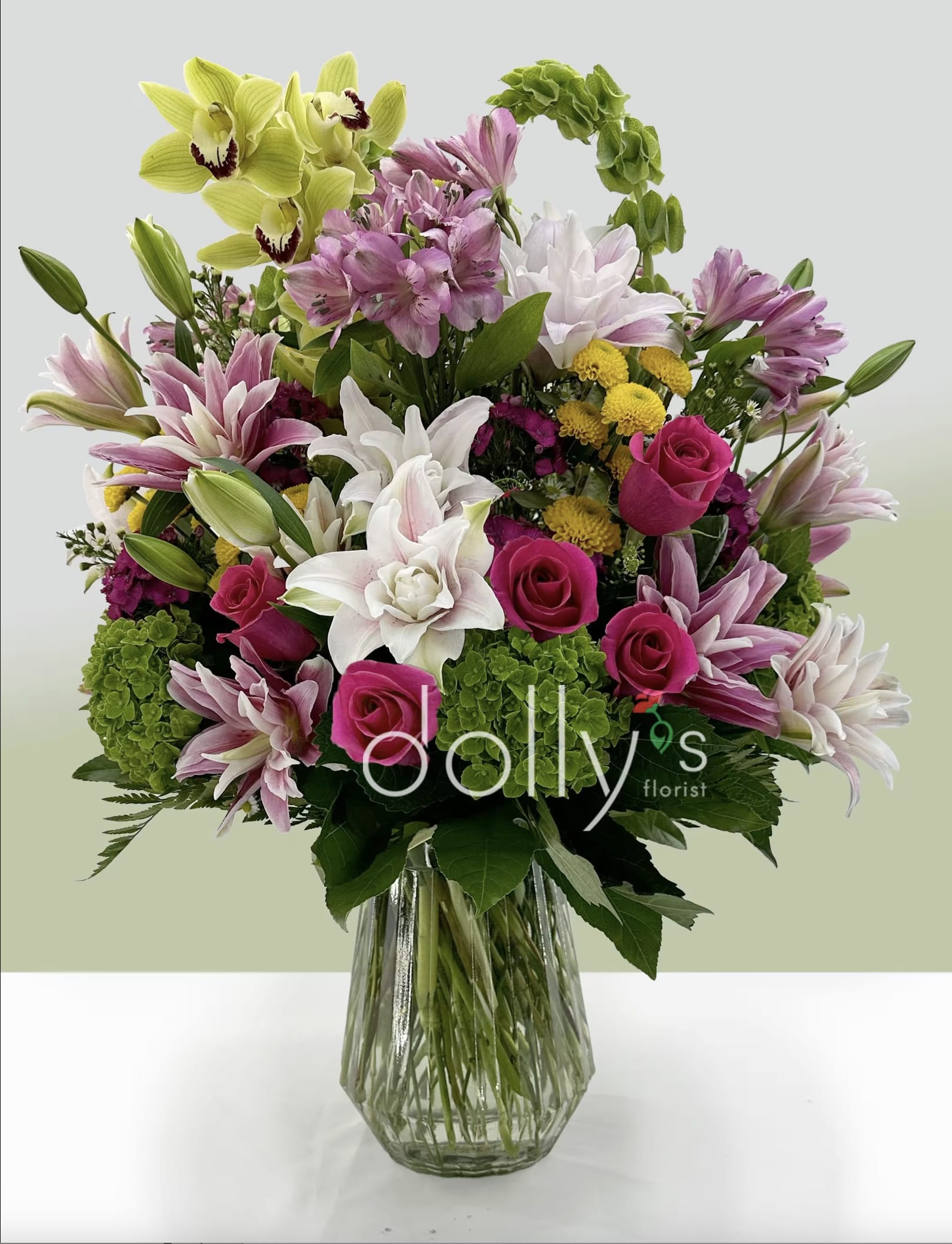Delilah's Garden - Hot pink roses and pale green orchids steal the show in this joyful flower arrangement perfect for a birthday bouquet, anniversary roses alternative, or a general celebratory gift. Delilah's Garden greets the recipient with a lush, rounded silhouette-an inviting mix of romantic pinks, crisp whites, fresh greens, and sunny yellow accents that instantly feels like a bright, thoughtful surprise.  The design combines hot pink roses, white-and-pink lilies, pale green orchids, alstroemeria, and clusters of yellow button chrysanthemums, densely arranged for a full, layered look. Blooms extend outward from a clear glass vase to create a generous, abundant presentation that reads elegant and lively; textures range from the soft petals of roses and lilies to the architectural touch of orchids and the cheerful pompom look of the chrysanthemums.  Ideal for the person you want to make feel truly celebrated-whether marking a birthday, anniversary, or simply saying "I'm thinking of you"-this arrangement arrives ready to display in the included clear glass vase. For best longevity, refresh the water every two days and recut stems at an angle before placing them back in the vase.