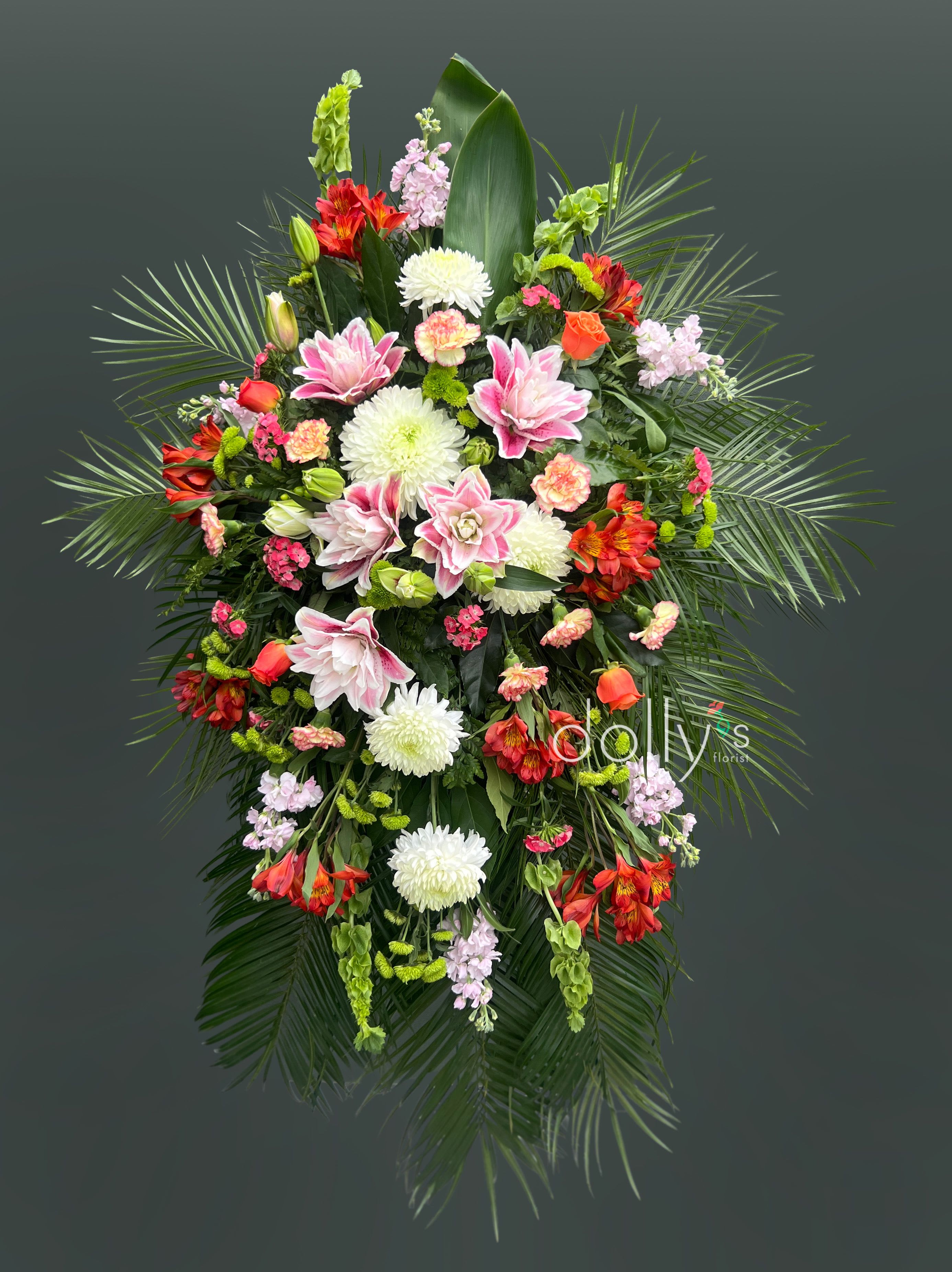 Dignity and Grace - A tall, formal standing spray that greets the room with quiet warmth-perfect for sympathy and funeral services. The vertical, symmetrical shape with sweeping palm fronds creates a dignified silhouette so the person you're honoring will be remembered with grace.  White chrysanthemums and soft pink blooms form a comforting center while orange-red alstroemeria, carnations, roses, and snapdragons add lively color and texture. Dark green foliage and broad tropical leaves frame the piece, giving strong contrast and a structured, ceremonial feel.  Designed to stand beside a casket or at a memorial space, this tribute suits intimate gatherings and larger services alike. Size and exact flowers may vary with seasonal availability.