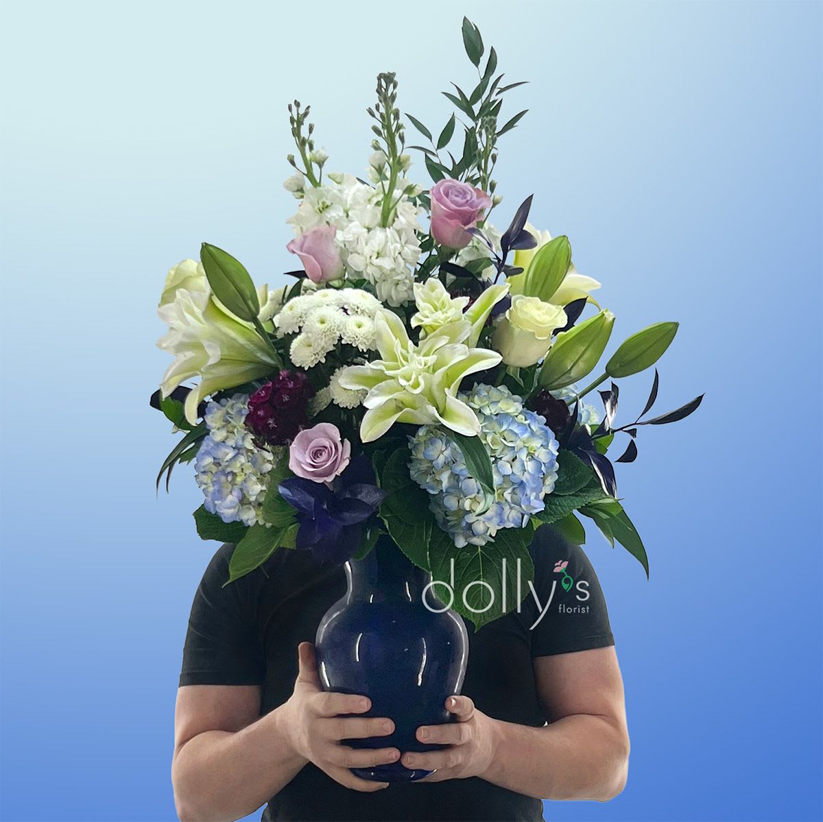 Blue Sky - Pale blue hydrangeas and large white lilies create a serene, elegant centerpiece that feels both refined and unexpectedly modern - perfect as a birthday surprise, a "just because" pick-me-up, or a romantic gesture. The arrangement's cool palette of white, lavender, blue, purple, pink, and green reads calm and celebratory; the vertical silhouette gives it a quietly dramatic presence that will make the recipient feel truly seen.  Rounded clusters of blue hydrangeas sit low and full while tall white lilies and purple chrysanthemums (with touches of stock and small white filler blooms) lift the eye. Dark foliage and bright blue decorative leaves add contrast; roses and other seasonal blooms weave gentle pink and lavender notes through the design. Everything is arranged in a substantial blue glass vase that anchors the composition and suits a prominent display on a dining table or entry console.  Thoughtful for Father's Day without feeling seasonal, this arrangement also works beautifully for anniversaries or sympathy where a refined, composed look is wanted. The substantial, upright styling holds its shape on arrival - trim stems occasionally and change the water to extend freshness, and choose the size that best fits the space you're gifting for.