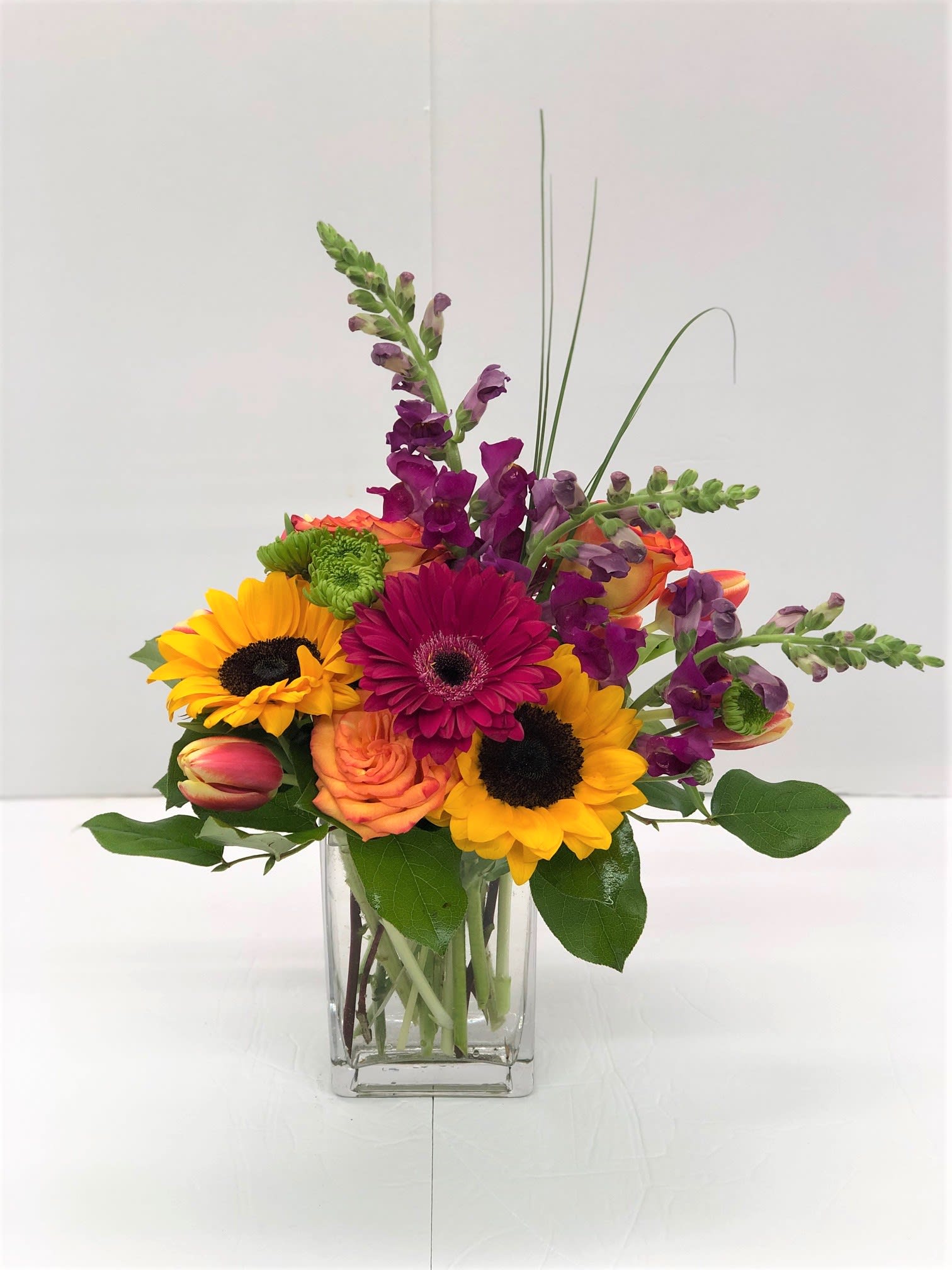 Mixed bouquet in a clear glass vase with sunflowers, roses, and purple spikes