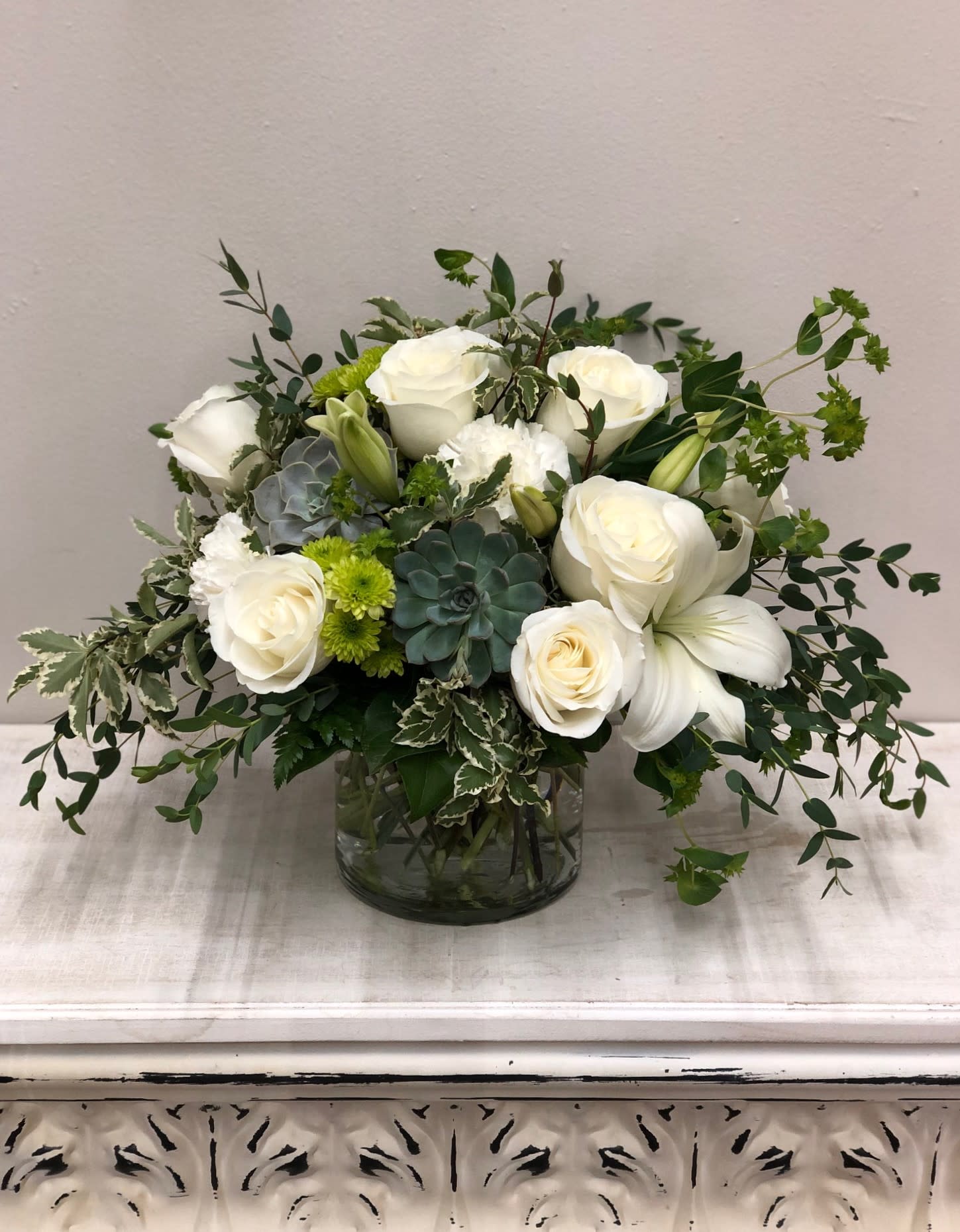 White roses and lilies in a glass vase with green succulents and foliage