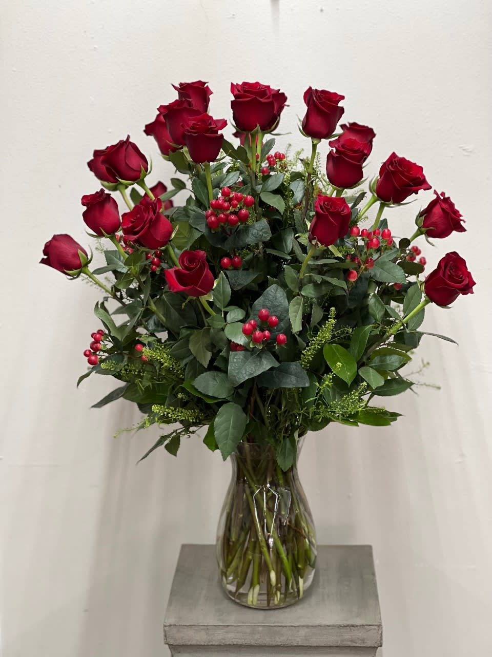 Tall arrangement of long-stem red roses with red berries in a clear glass vase
