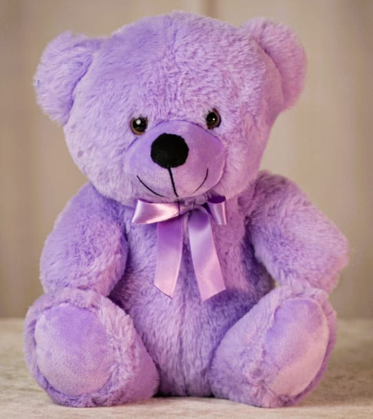 Lavender plush teddy bear with a satin bow sitting against a neutral background.