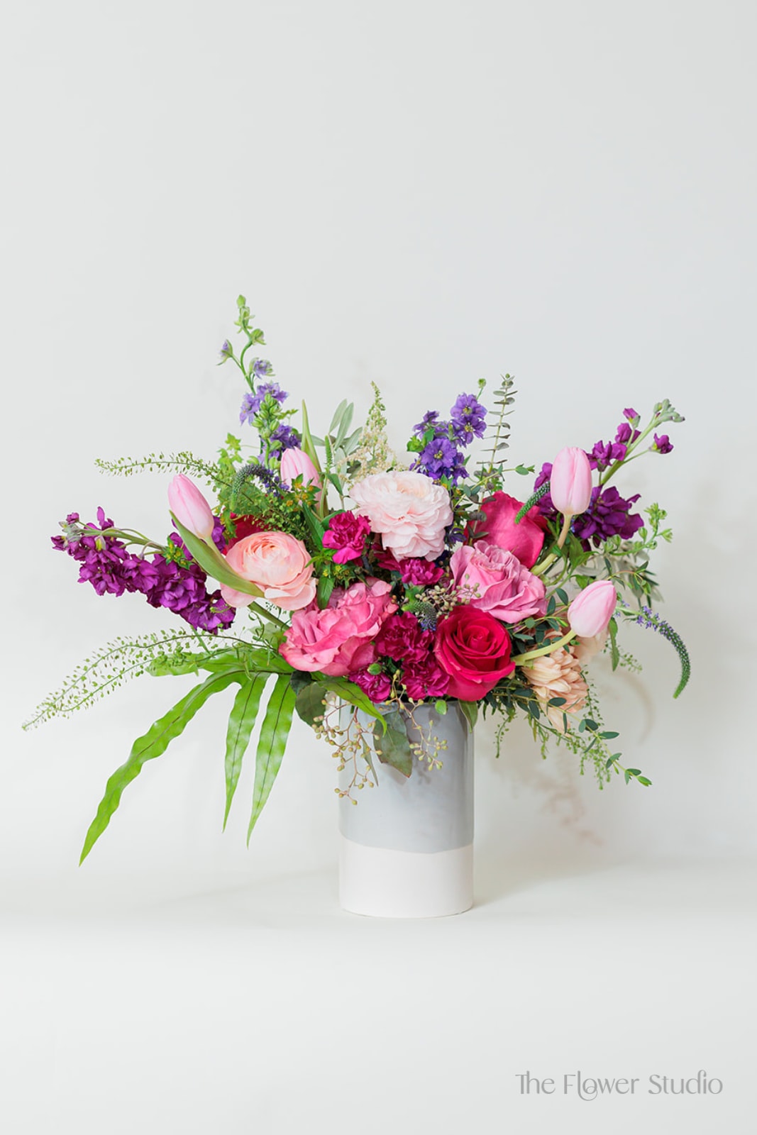 Asymmetrical pink and purple flower arrangement with tulips and roses in a tall white and gray cylinder vase