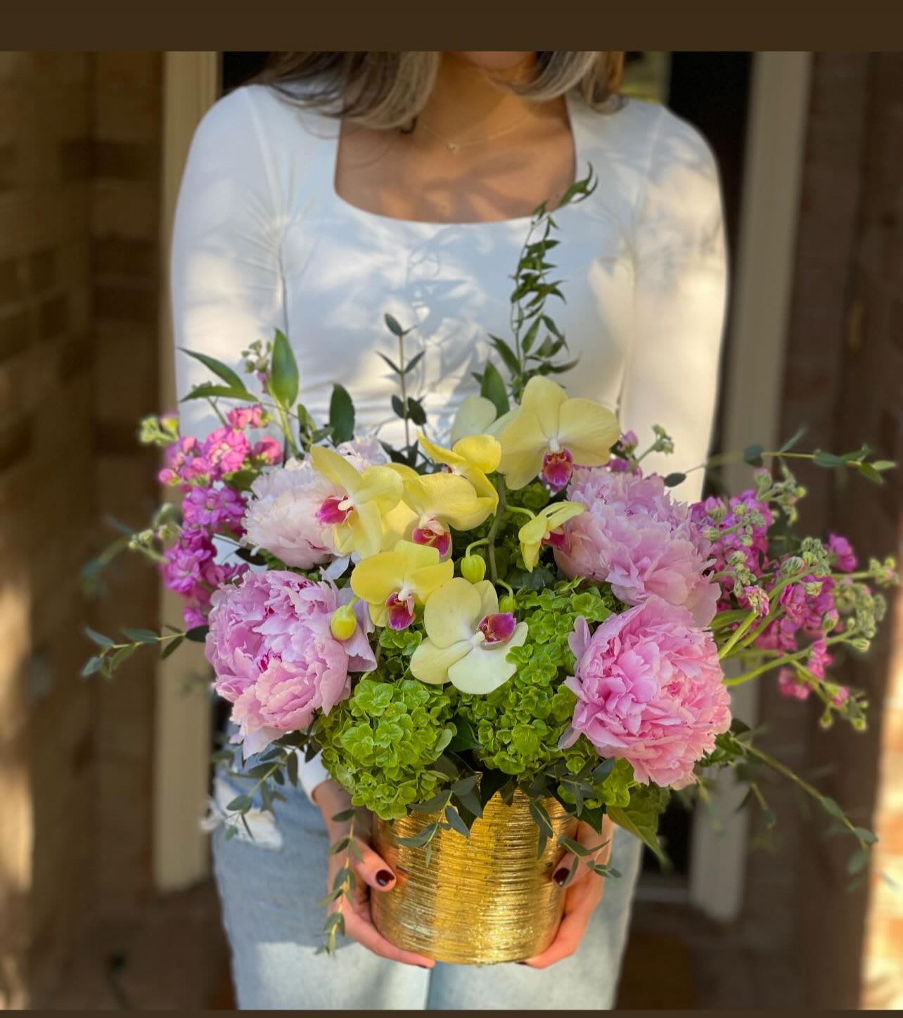 Lush arrangement of pink peonies, yellow orchids and green hydrangeas in a gold pot held by a person in white.