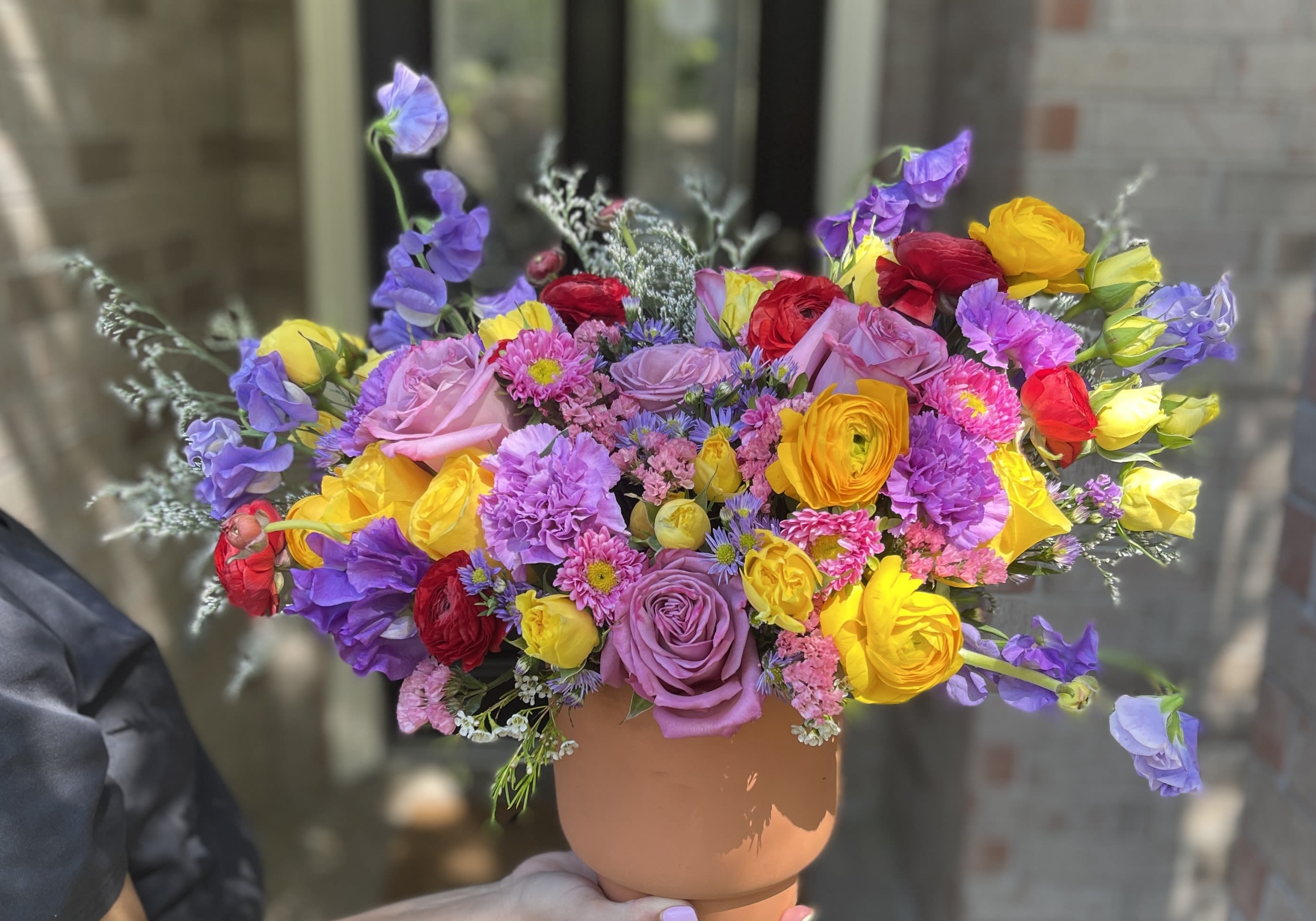 Colorful bouquet of roses, ranunculus, and purple blooms in a tan vase