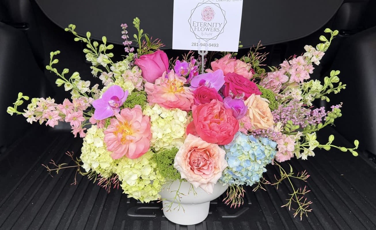 Amy - A lush, garden-style centerpiece that reads like everyday luxury-Amy is a large, low flower arrangement perfect as a birthday bouquet, anniversary gift, or spring celebration. Soft pink roses and blushing peonies take center stage so the person you're thinking of will feel cherished, while sprays of Phalaenopsis orchids and delicate branching blooms add graceful height and movement for a romantic, joyful presence.  Crafted in a clean white ceramic vase with a small floral pick sign, this composition mixes pastel lavender, peach, white and pale blue tones with brighter pink accents for cheerful contrast. Hydrangeas and assorted filler greenery fill the wide, abundant silhouette; the piece is designed to sit low on a table and make an immediate visual impact without overwhelming the space.  Ideal for celebrating milestones, saying thank you, or brightening someone's day, Amy arrives ready to admire-no arranging required. Keep the vase topped up with fresh water and trim stems as needed to extend bloom life; its generous scale makes it a striking gift for someone you want to make feel truly seen.