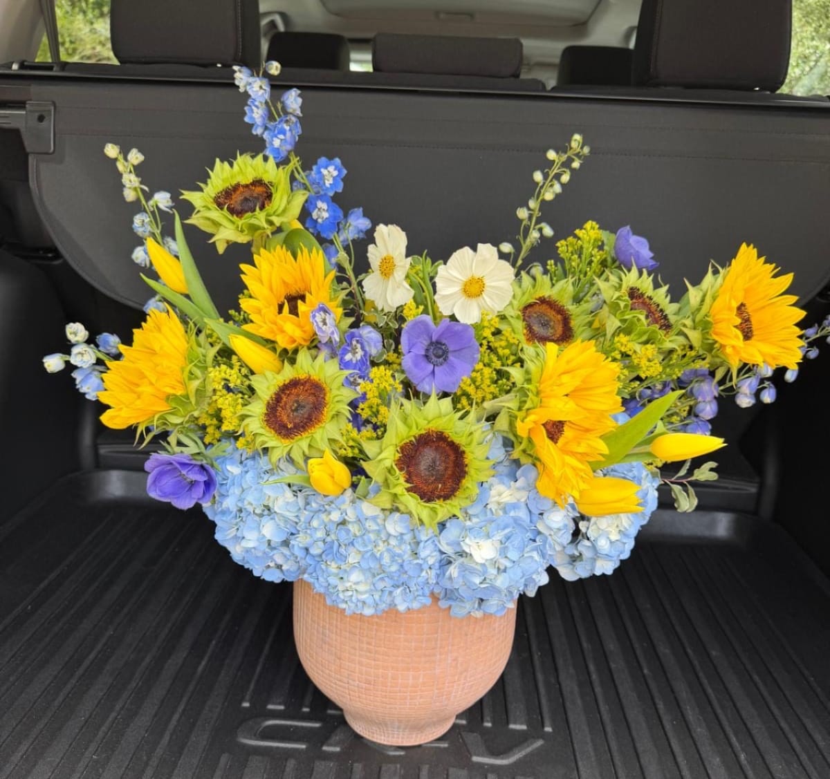Alba Sun - Bright yellow sunflowers take center stage in this cheerful mixed-color flower arrangement, making it an ideal birthday bouquet, congratulations gift, or springtime surprise. The wide, garden-style silhouette-tall stems rising above a lush base-creates lively movement so the person you're thinking of will feel genuinely celebrated the moment it arrives.  A dense collar of pale blue hydrangeas forms the cool counterpoint to the sunflowers, while purple and white accents-anemones, delphinium touches, spring tulip  and gentle contrast. The design sits in a ceramic vase that gives the arrangement a substantial, decorative presence and modern, slightly rustic charm; the composition is large, airy, and abundantly rounded to read beautifully on a dining table or entry console.  Perfect for birthdays, congratulations, or a bright "thinking of you" gesture, this arrangement arrives ready to display in its ceramic vessel. Its full shape and durable flowers make it especially suitable for gifting; for best enjoyment, keep in fresh water, away from direct heat, and refresh the vase water every couple of days.