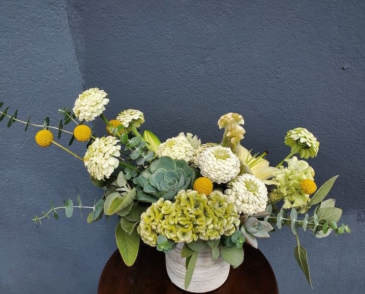 Low arrangement of white blooms, green succulents, and yellow accents in a white pot against a dark wall