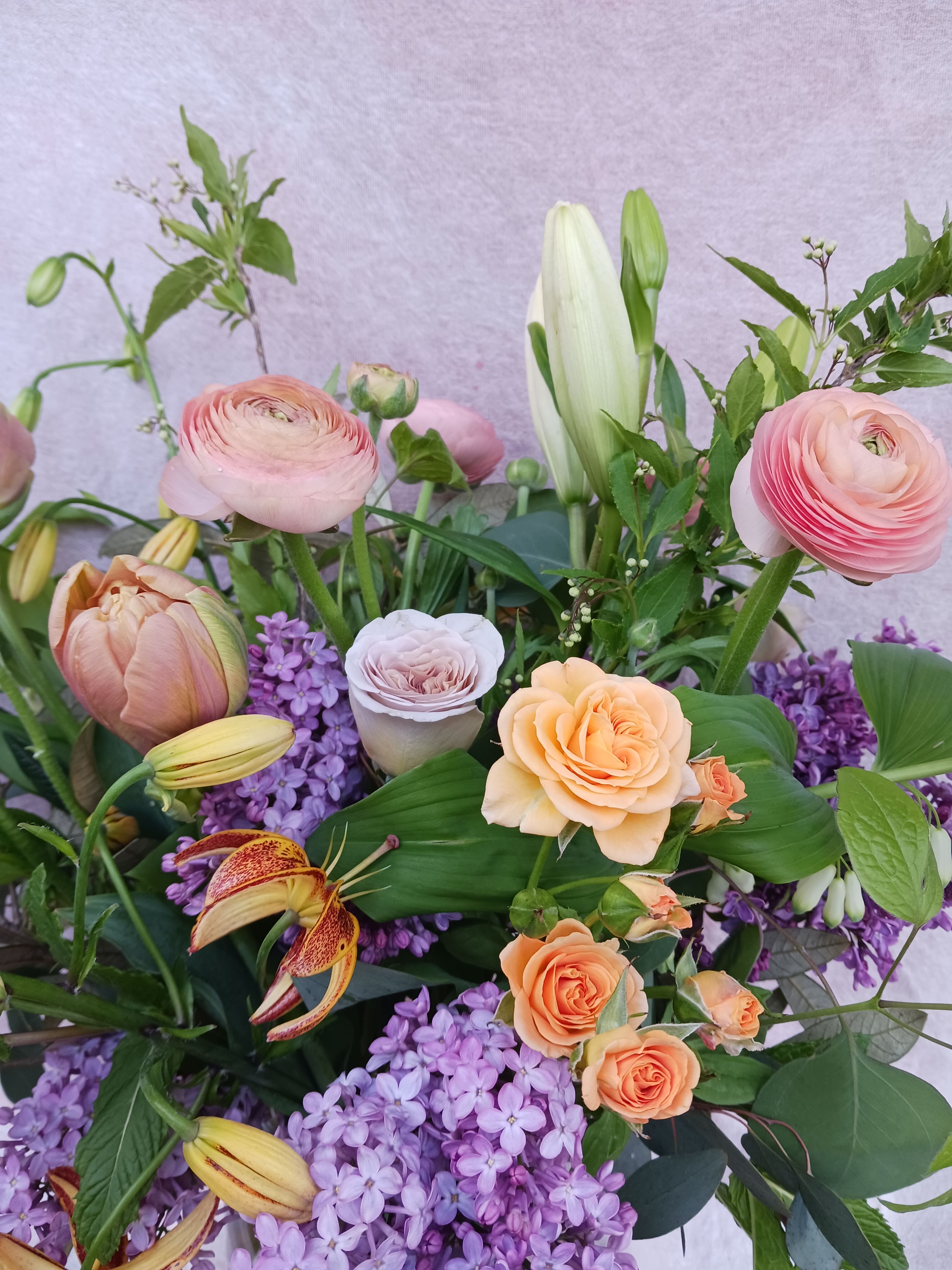 Garden-style arrangement with peach roses, pink ranunculus, lilies, and purple lilacs