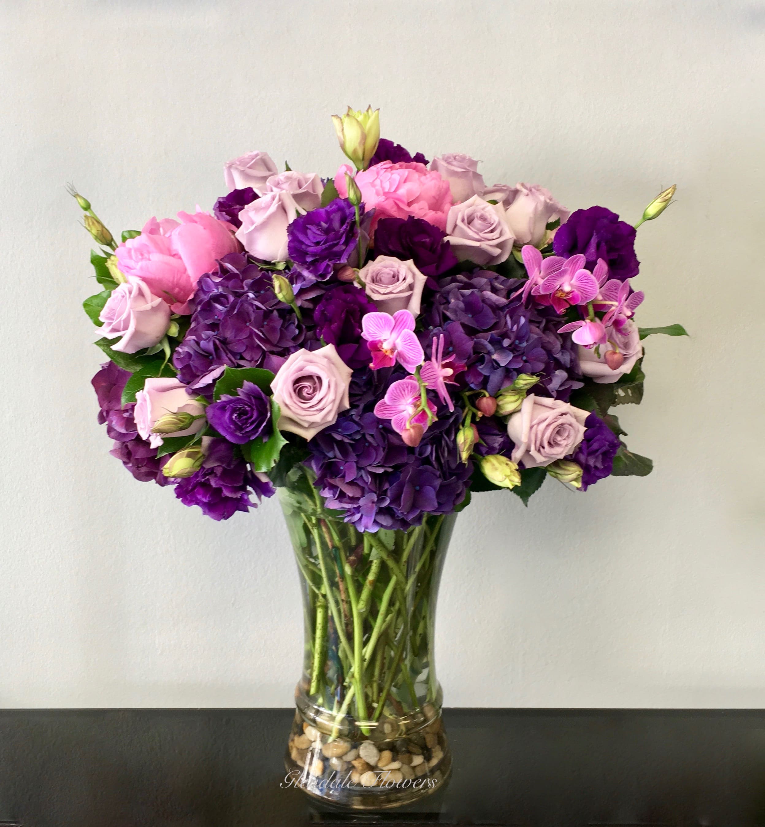 Tall glass vase of purple hydrangeas, lavender roses, and pink blooms in a rounded arrangement