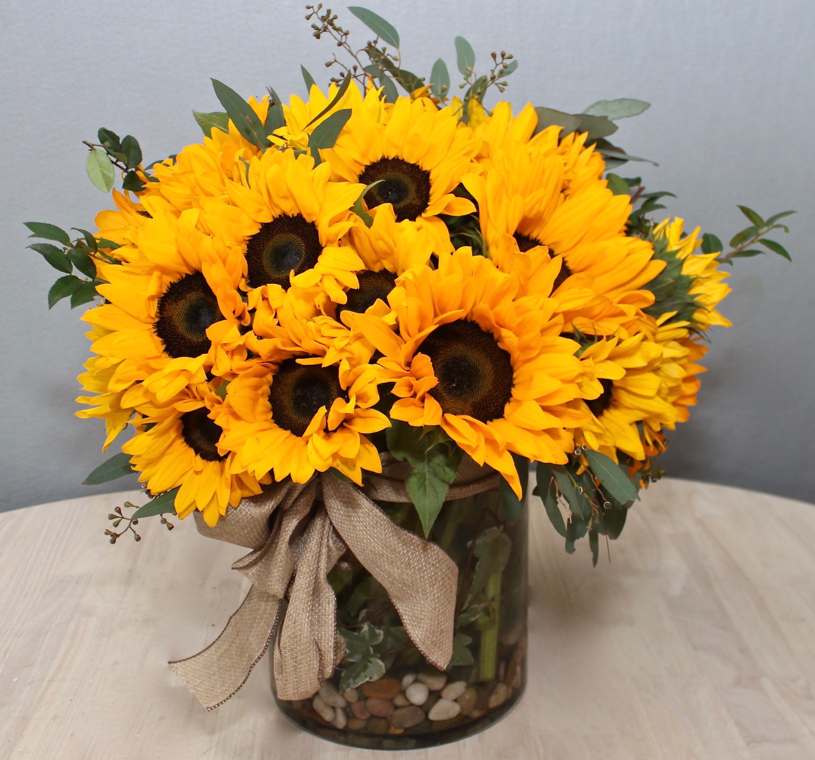Low round bouquet of yellow sunflowers in a clear glass vase with stones and a burlap bow