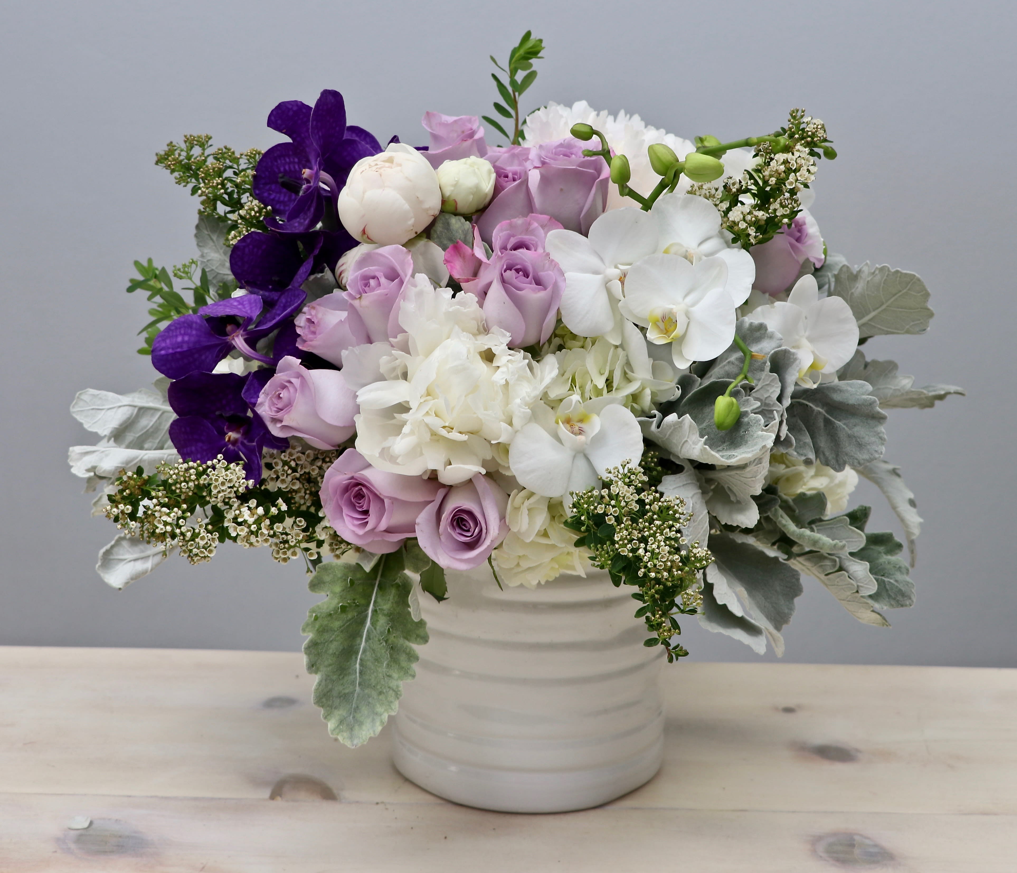 Low white vase filled with lavender roses, white hydrangeas, and orchids with purple accents