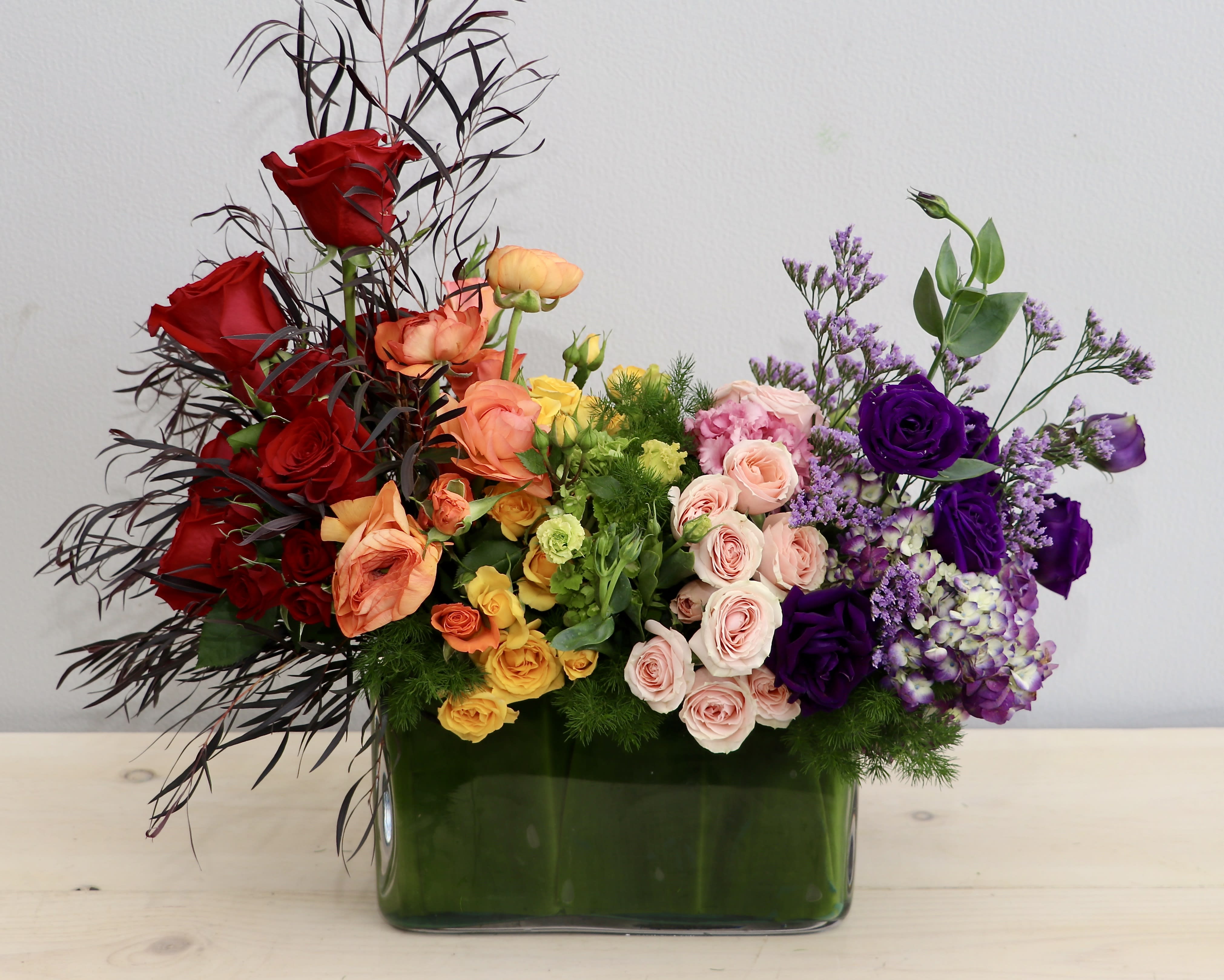 Low rectangular arrangement of red, yellow, peach, and purple flowers in a glass vase.