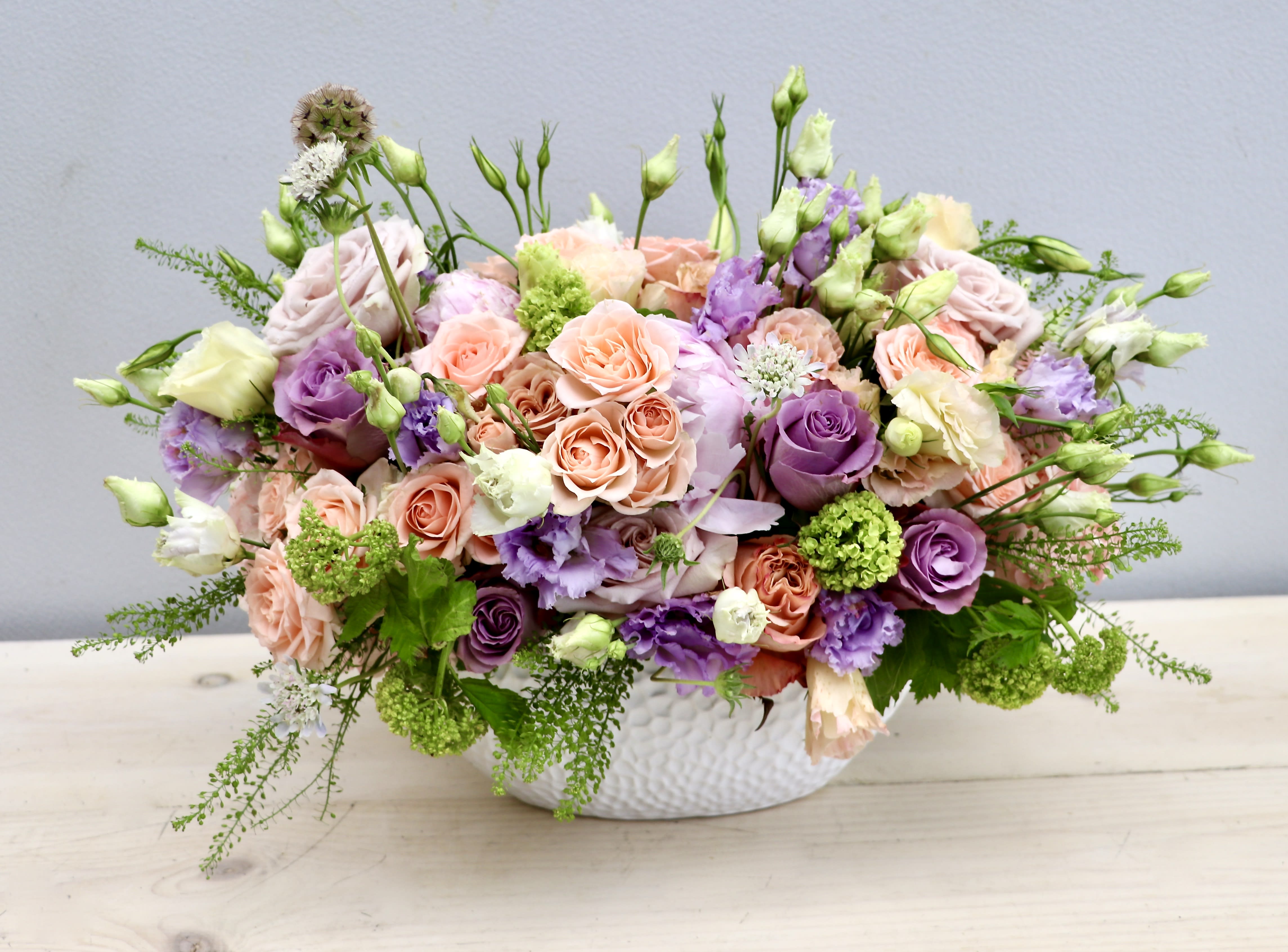 Low centerpiece with peach and lavender roses, lisianthus and green blooms in a white oval vase