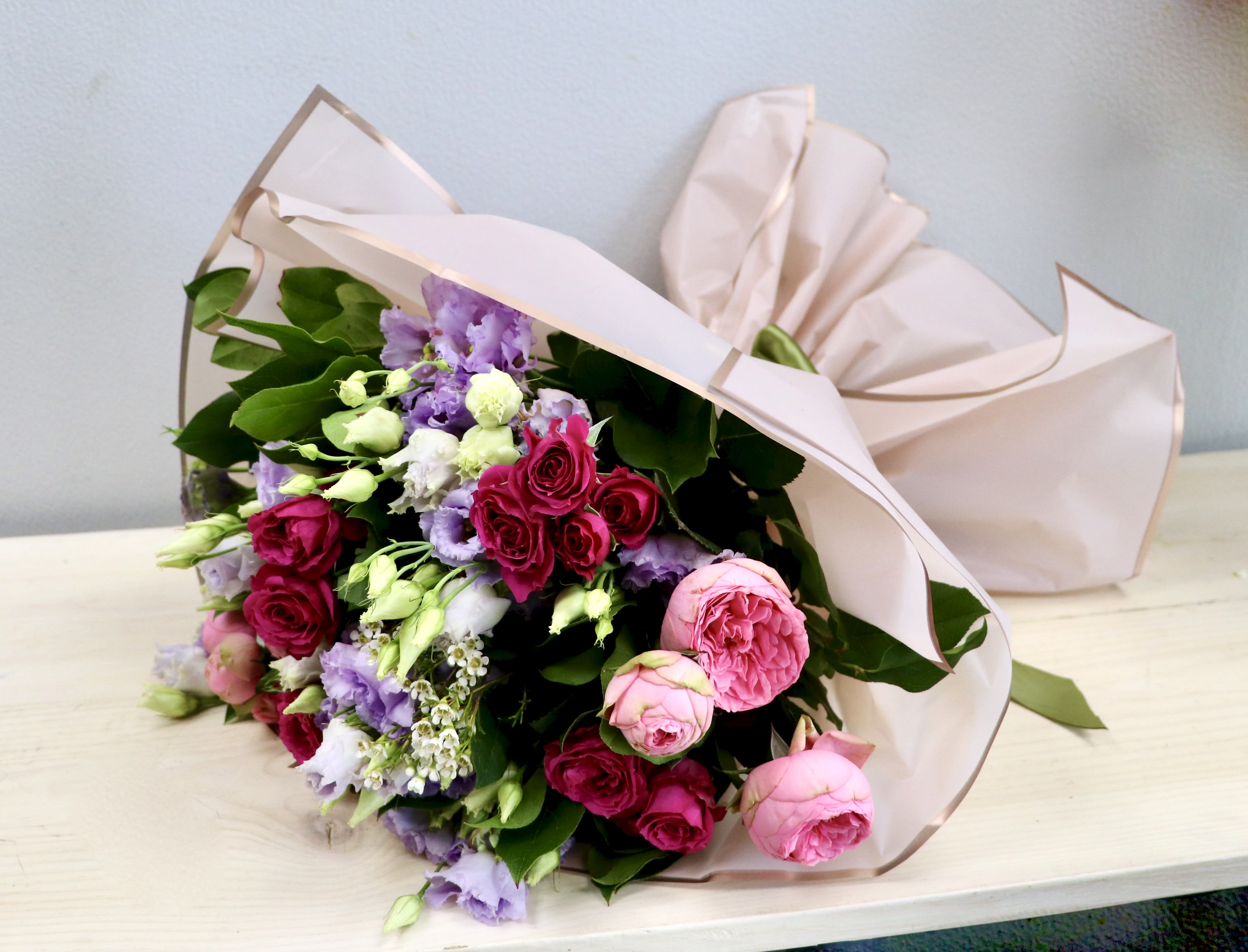 Pink and lavender bouquet of roses and lisianthus wrapped in blush paper, resting on a light wood surface