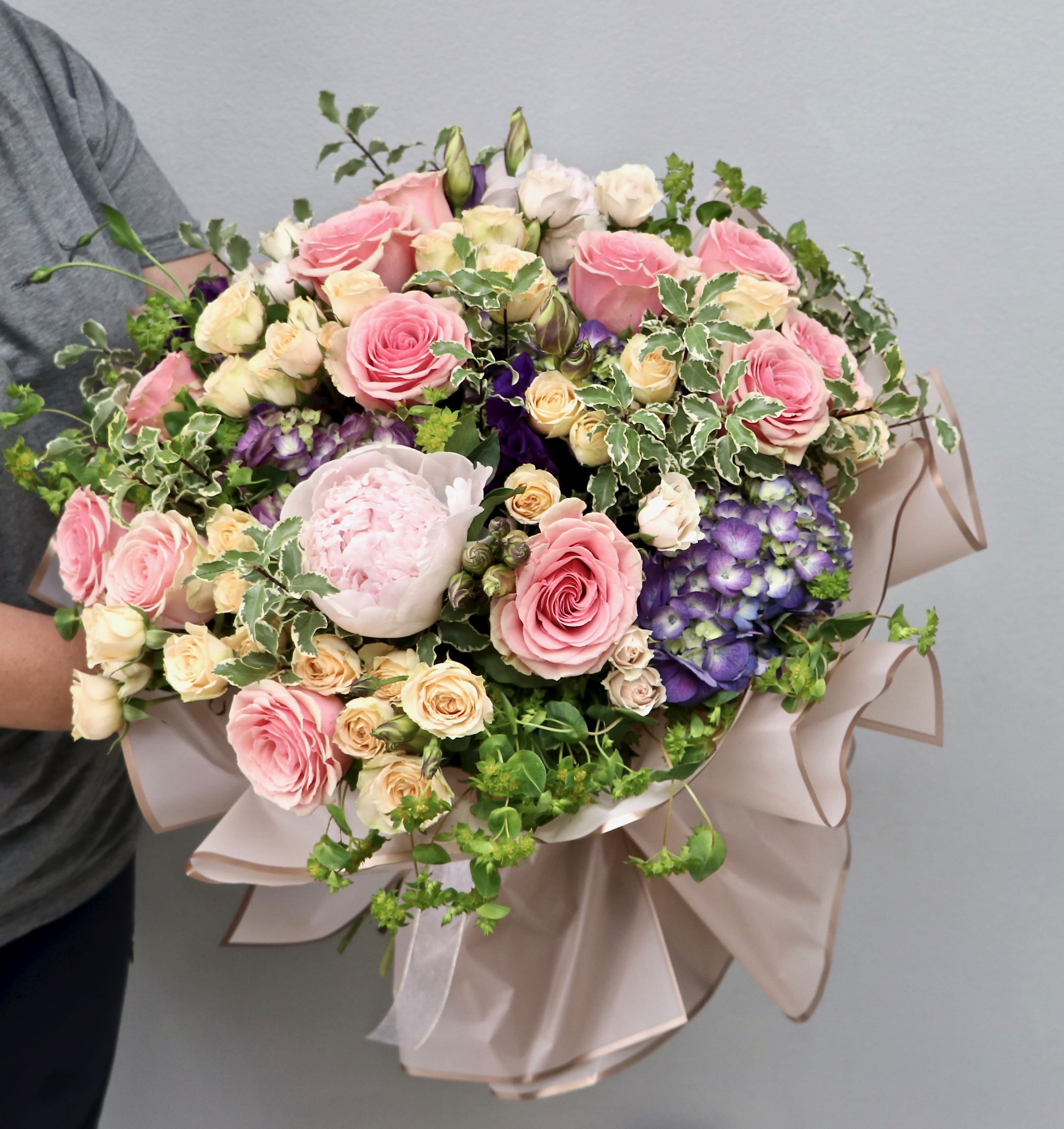 Large hand-tied bouquet of pink roses, cream flowers, and purple hydrangeas wrapped in blush paper