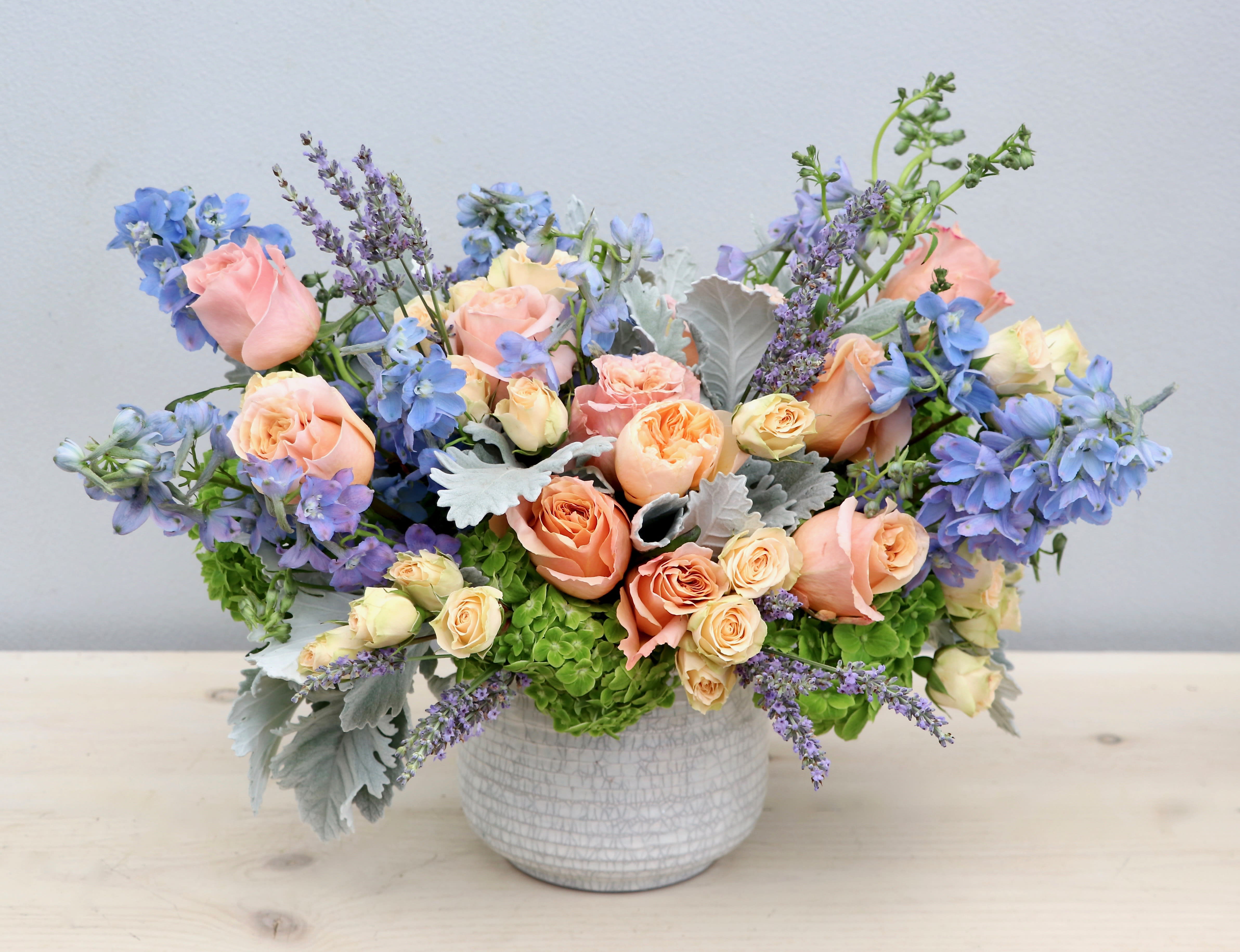 Low arrangement of peach roses and blue flowers in a white ceramic bowl vase
