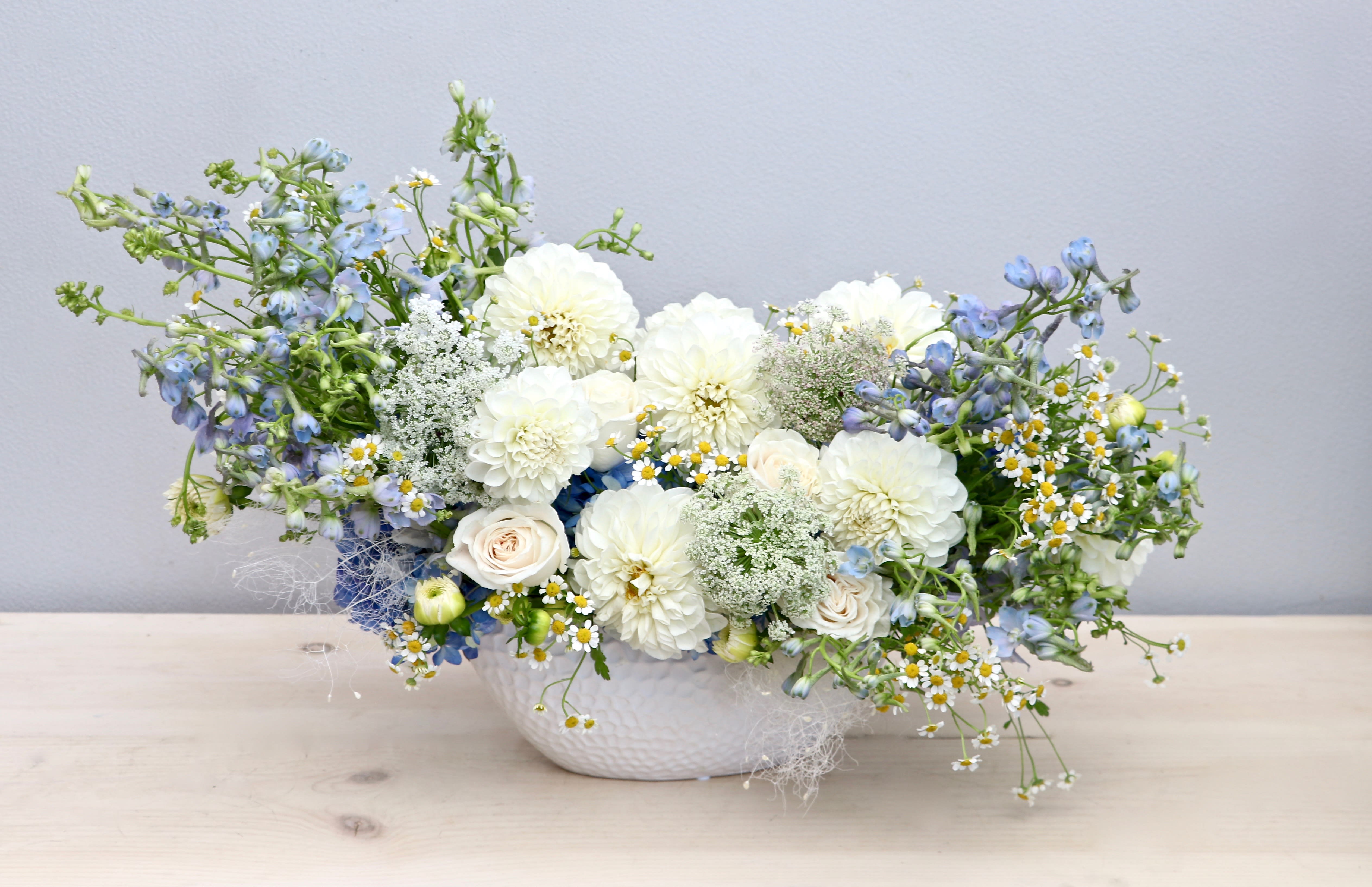 Low white ceramic vase with white dahlias, pale roses, blue delphinium, and small white daisies in an airy arrangement