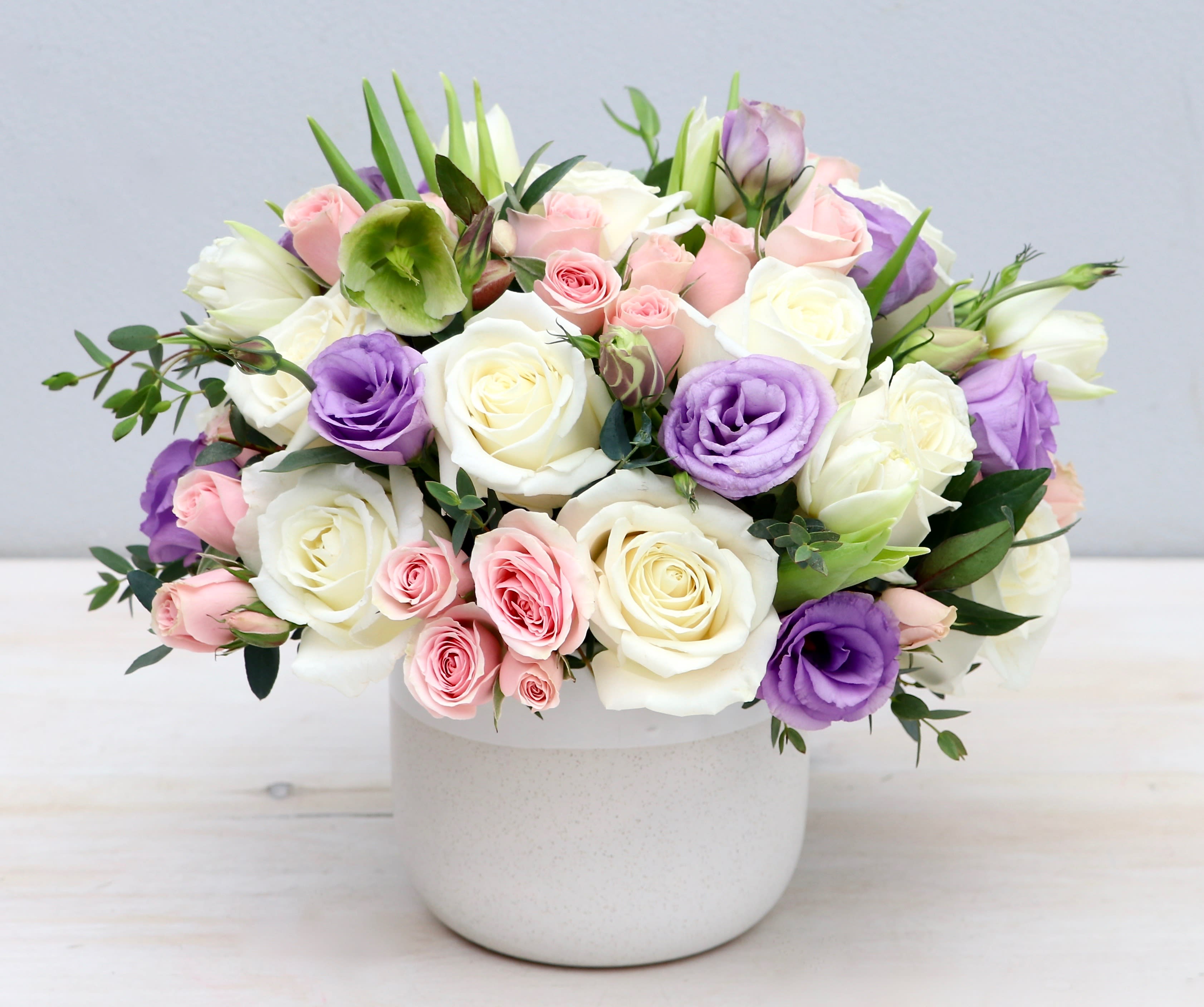 Round arrangement of white roses, pink roses, and lavender lisianthus in a white ceramic pot