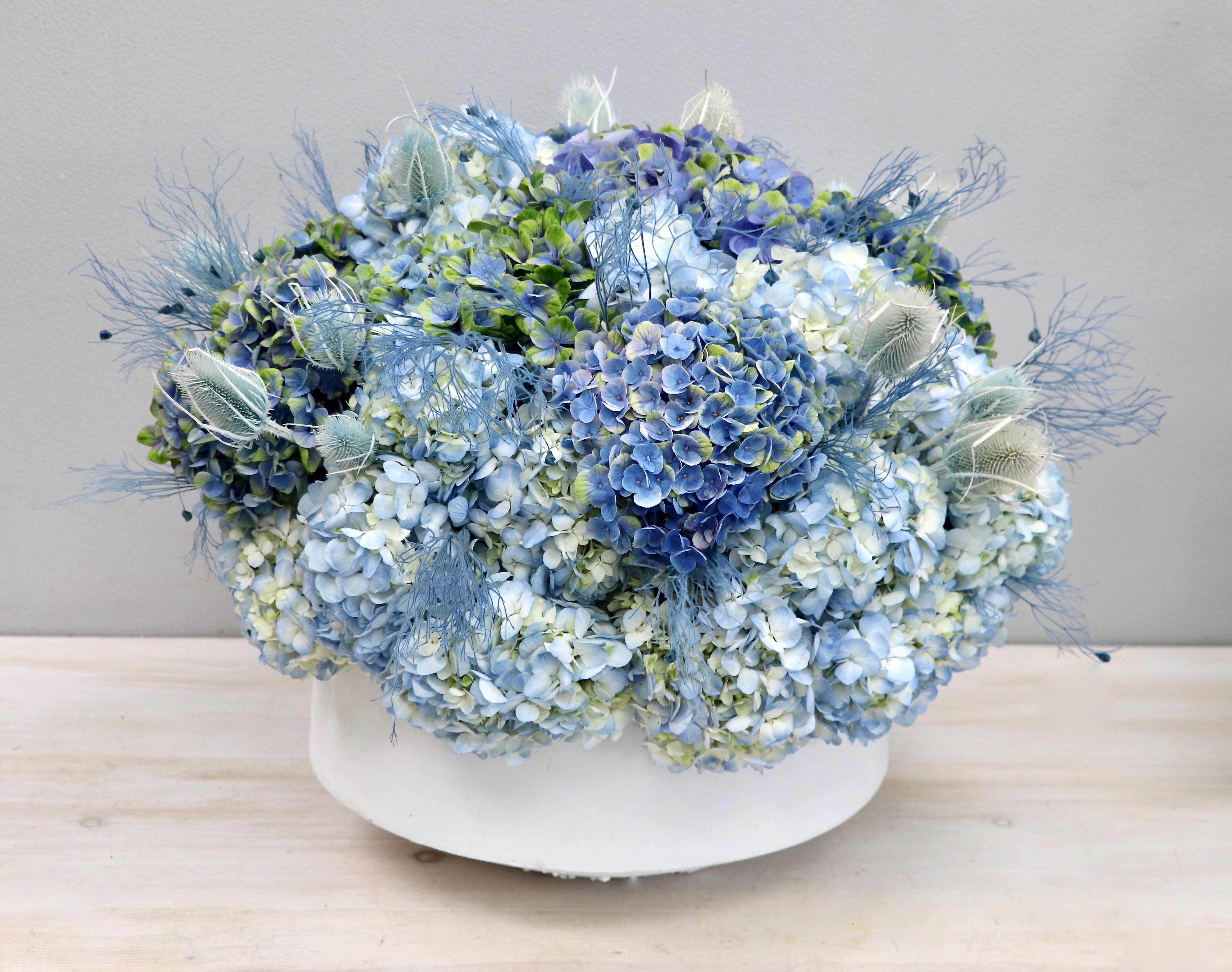 Low round arrangement of blue and white hydrangeas with airy accents in a white ceramic bowl.