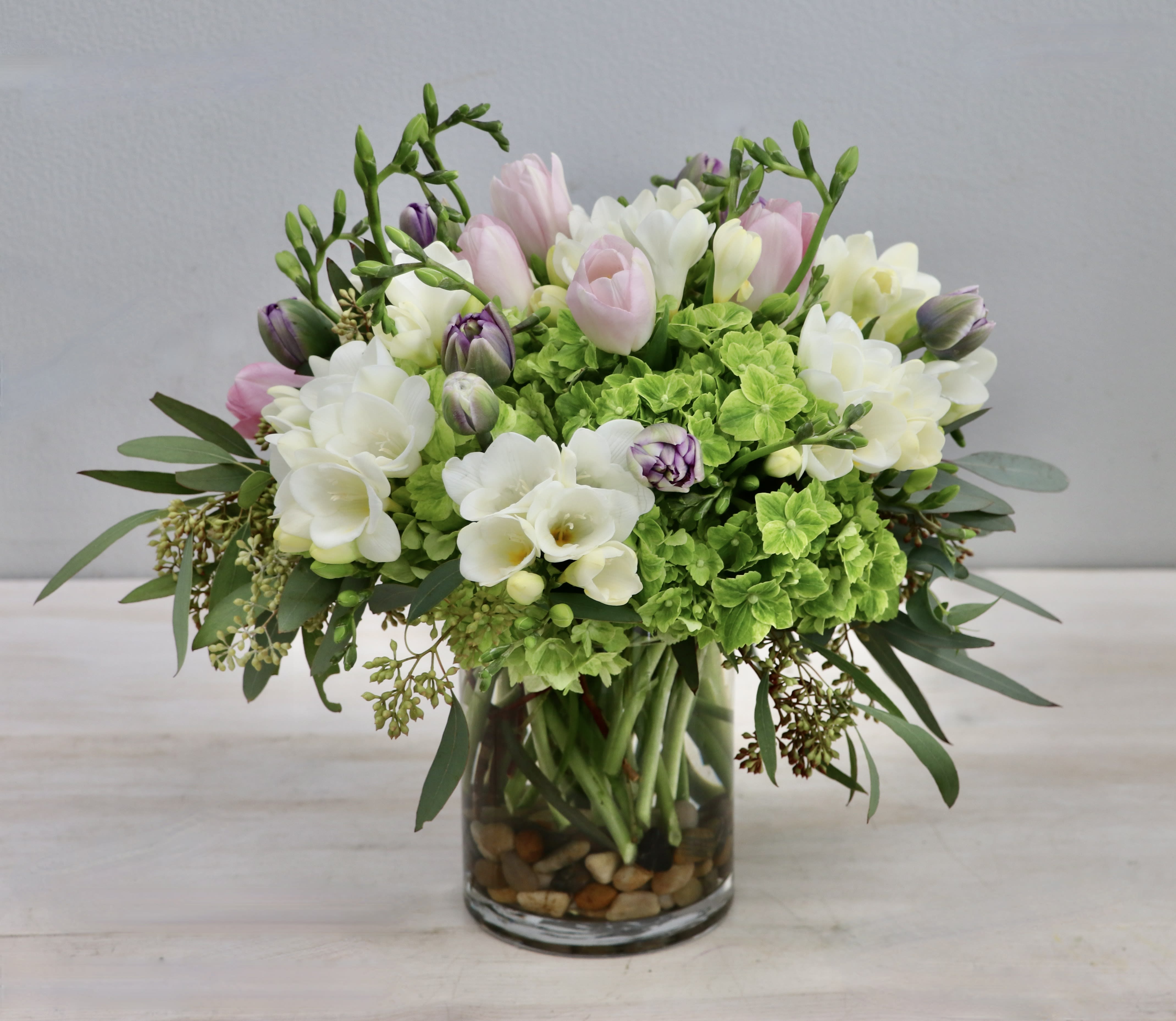 Compact arrangement of pink tulips, white freesias, and green hydrangeas in a clear glass vase with stones.