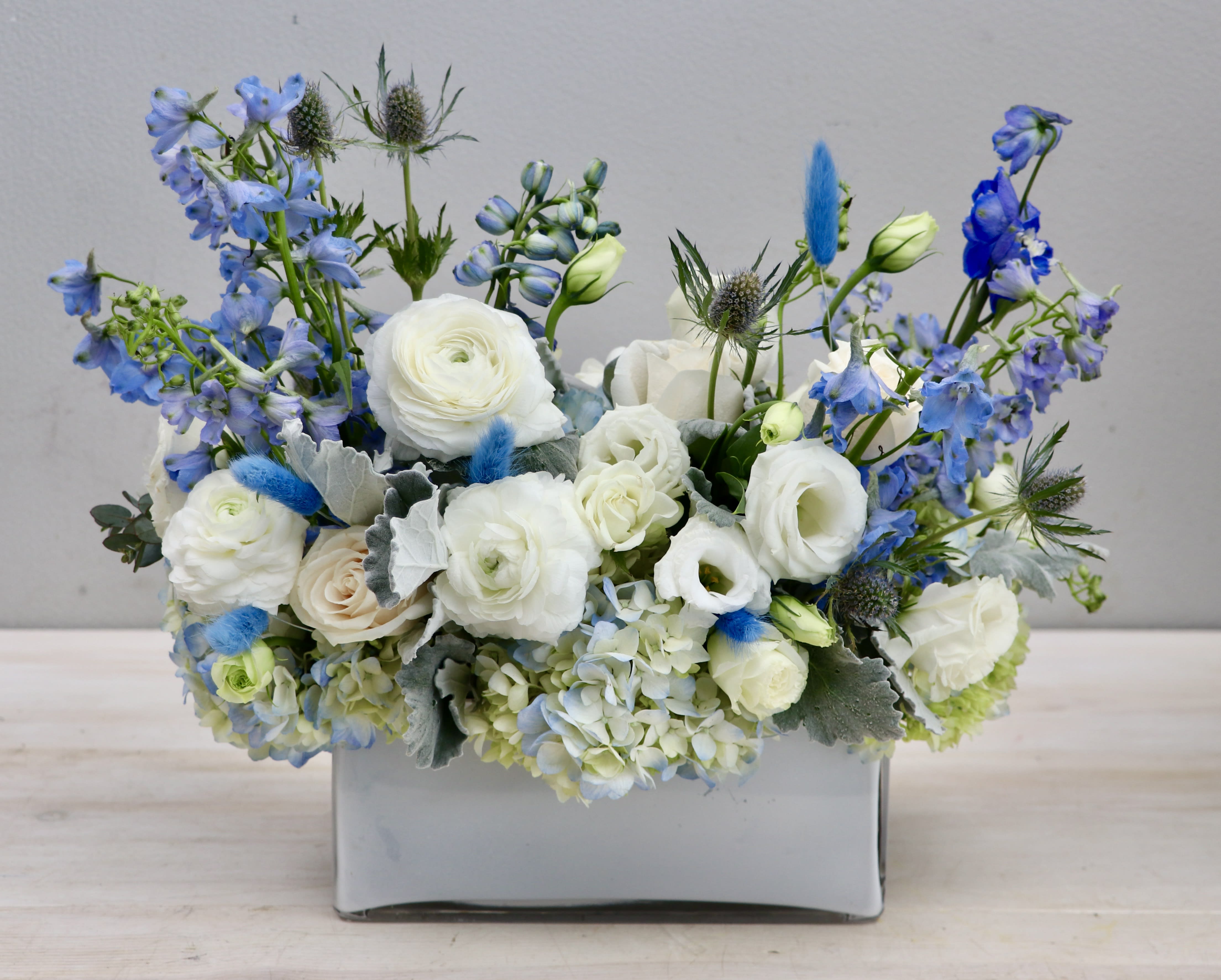 Low arrangement of white roses and ranunculus with blue delphinium in a gray ceramic vase