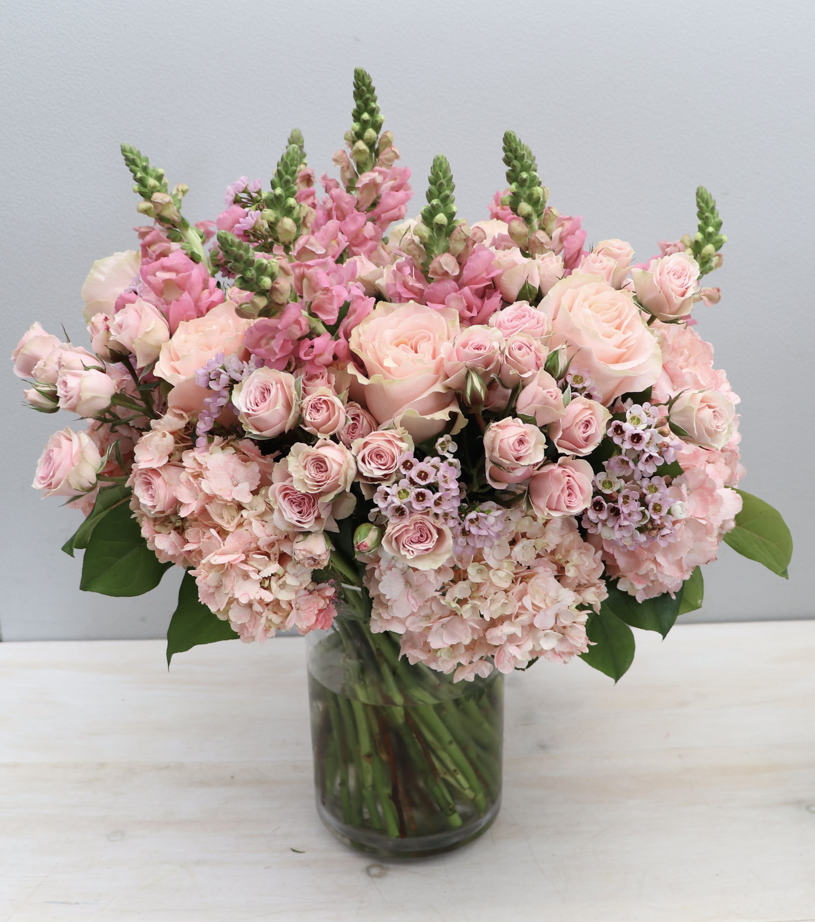 Large arrangement of soft pink roses, snapdragons, and hydrangeas in a clear glass vase.