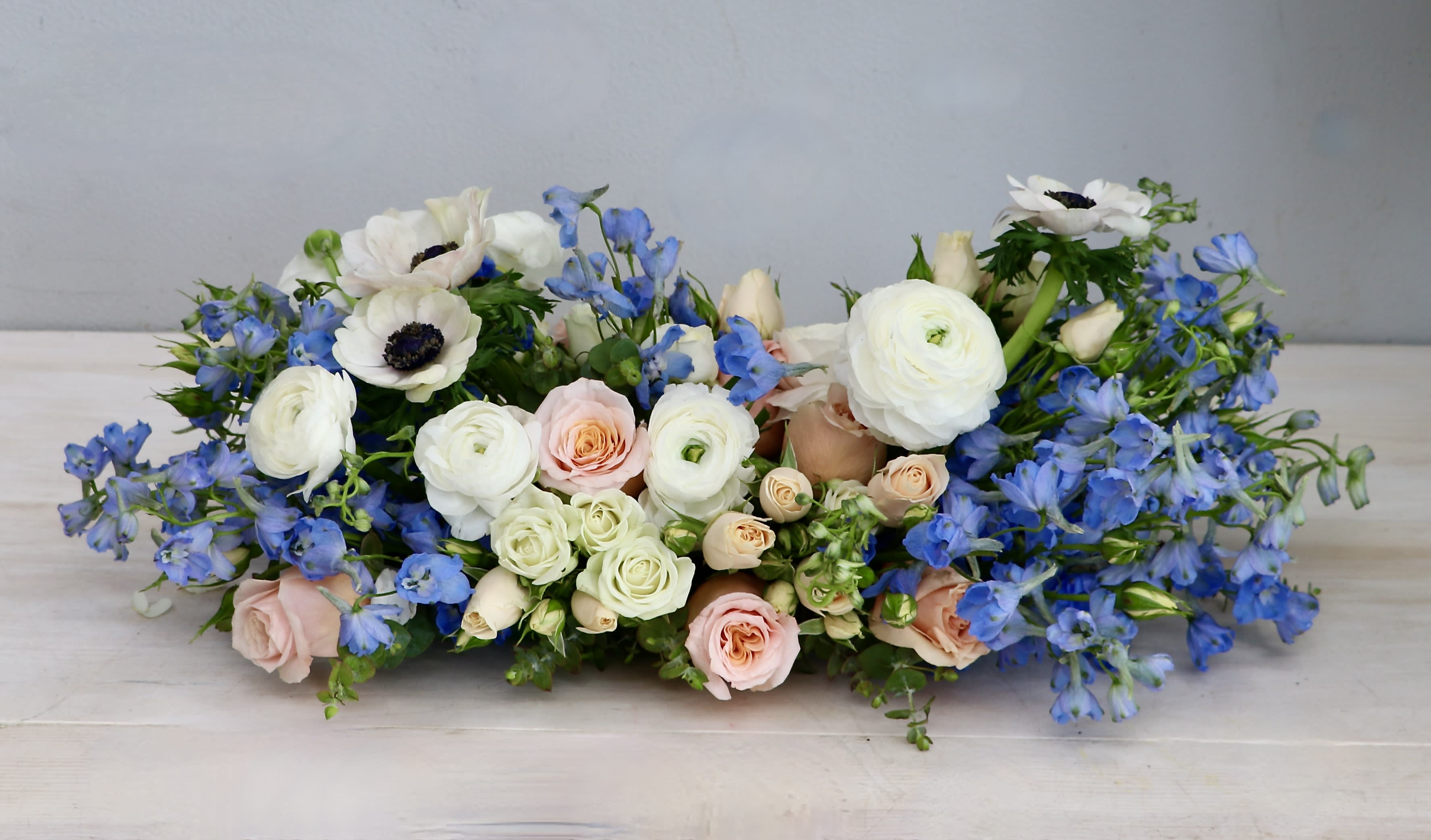Low centerpiece of white ranunculus, blue delphinium, and blush roses in a long horizontal arrangement