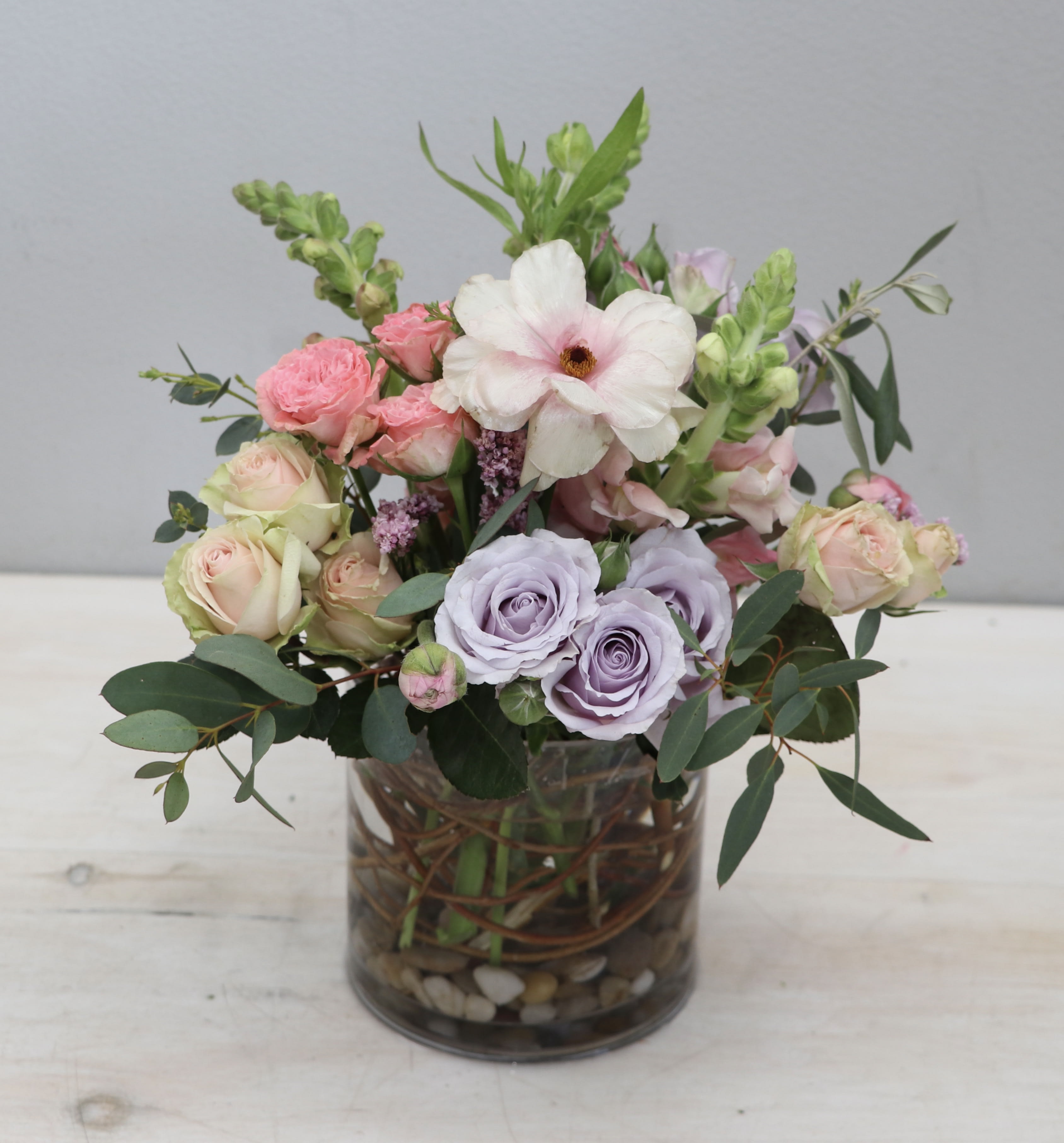 Pastel arrangement of pink and lavender roses, anemone, and snapdragons in a clear glass vase with stones