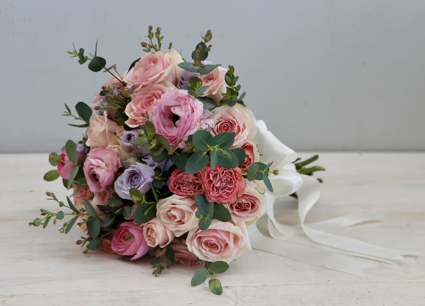 Pastel pink and peach rose bouquet with lavender blooms and white ribbon trailing on a light surface