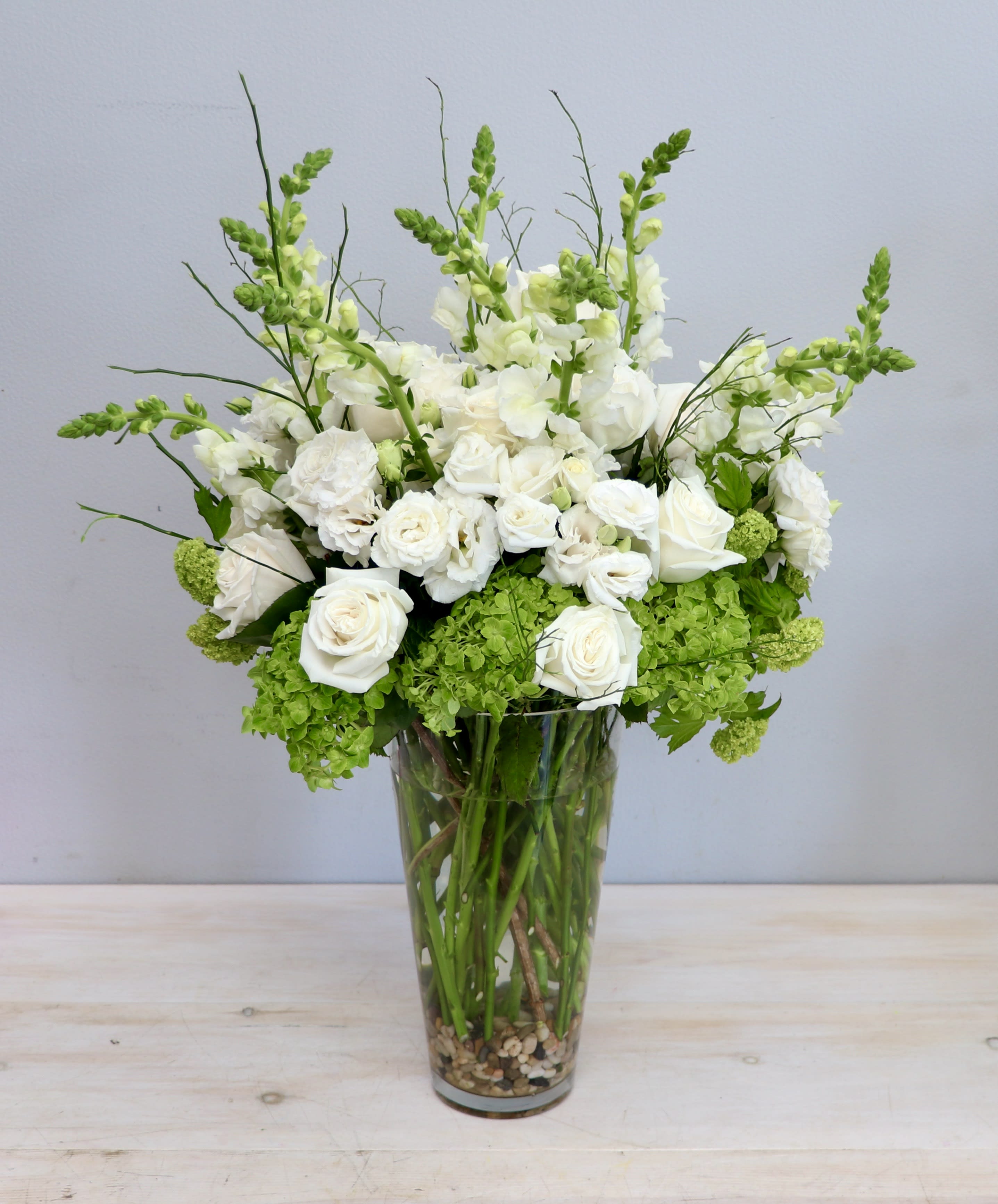 Tall arrangement of white roses and snapdragons with green blooms in a clear glass vase.