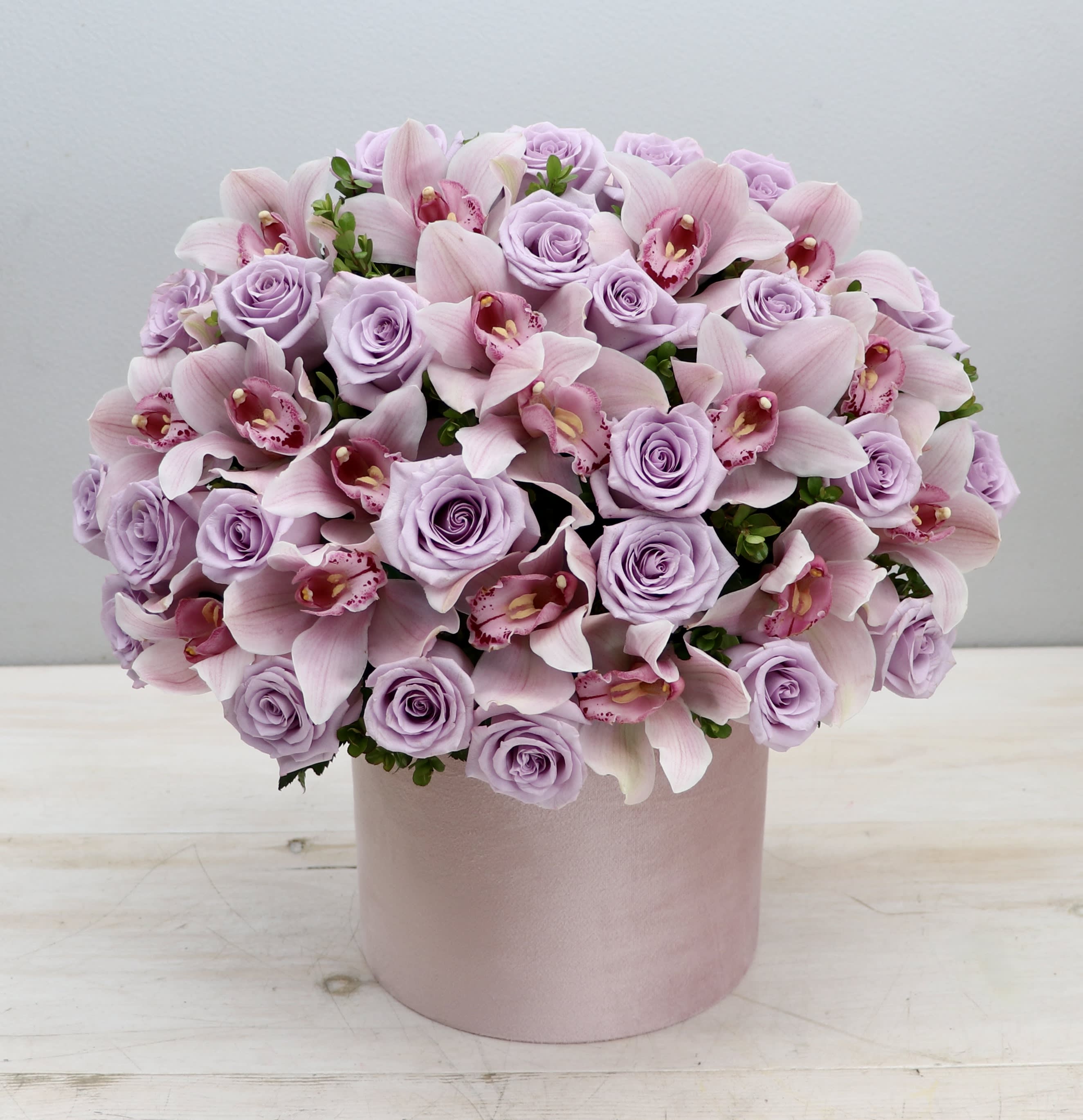 Round arrangement of pale lavender roses and pink orchids in a light pink hat box container.